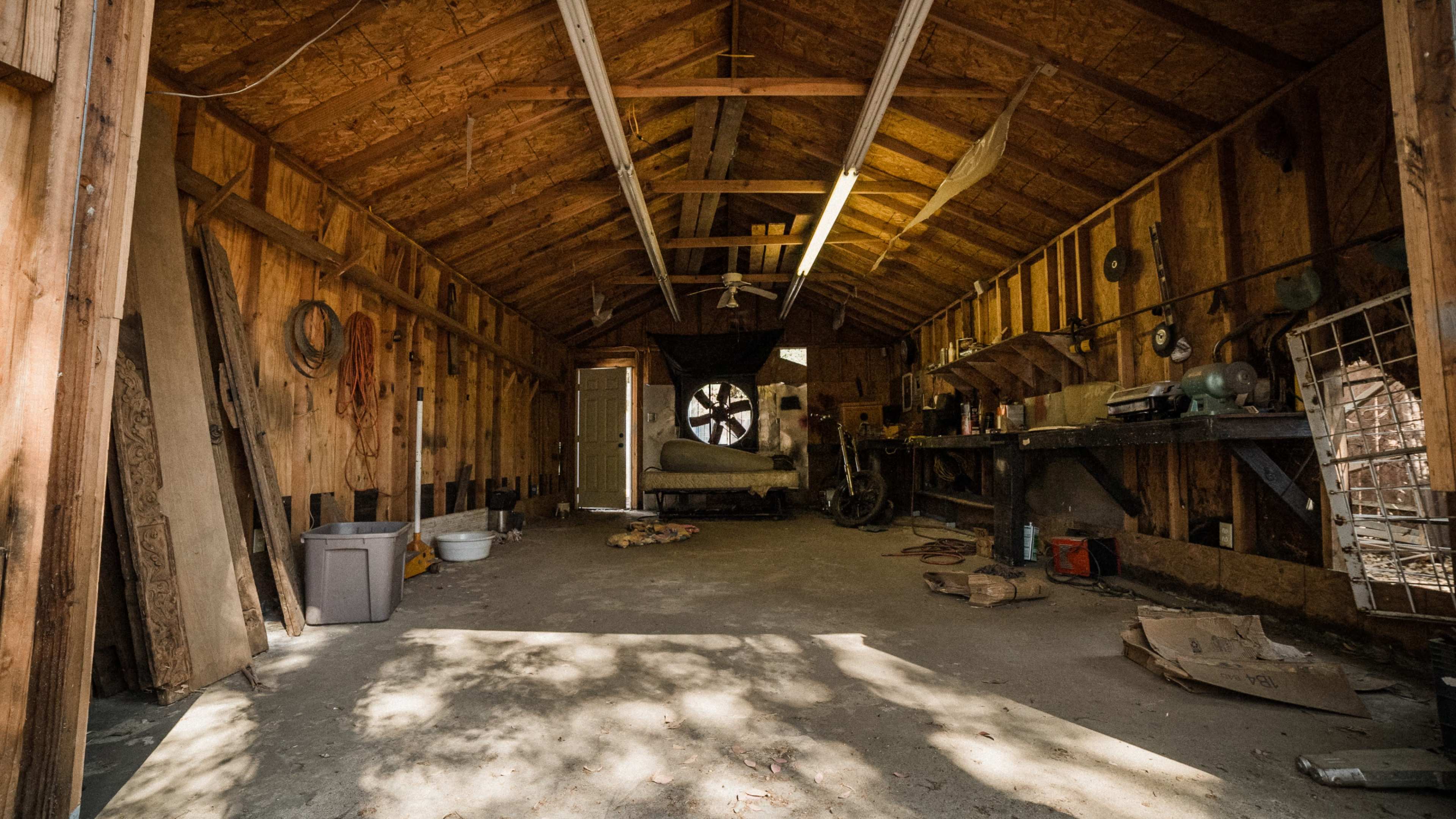 An empty wooden shed with a concrete floor, featuring exposed beams, tools on a workbench, and a large wheel in the back.