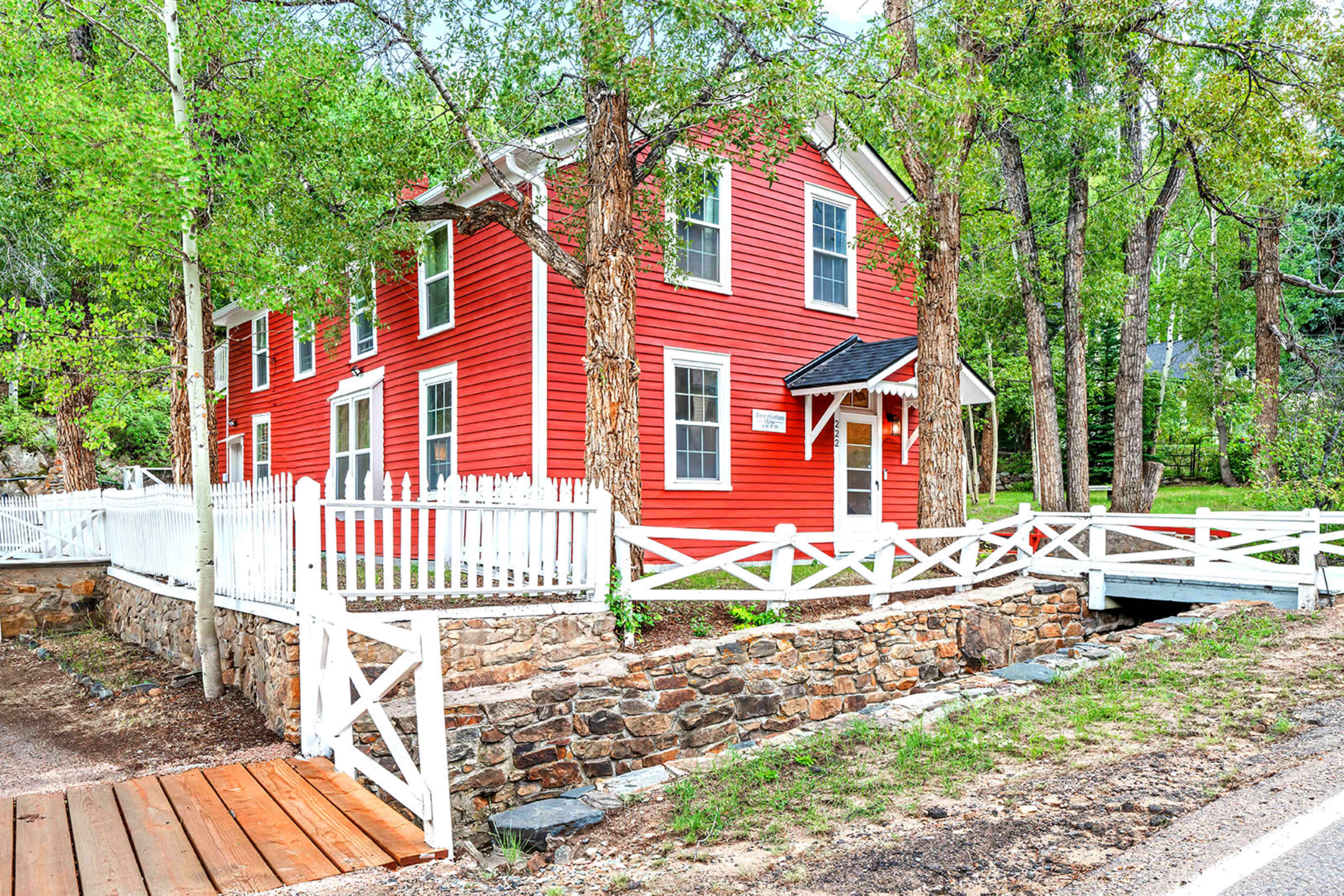 The image shows a red two-story house surrounded by trees and a white picket fence, with a stone wall and a small bridge leading to the front entrance.