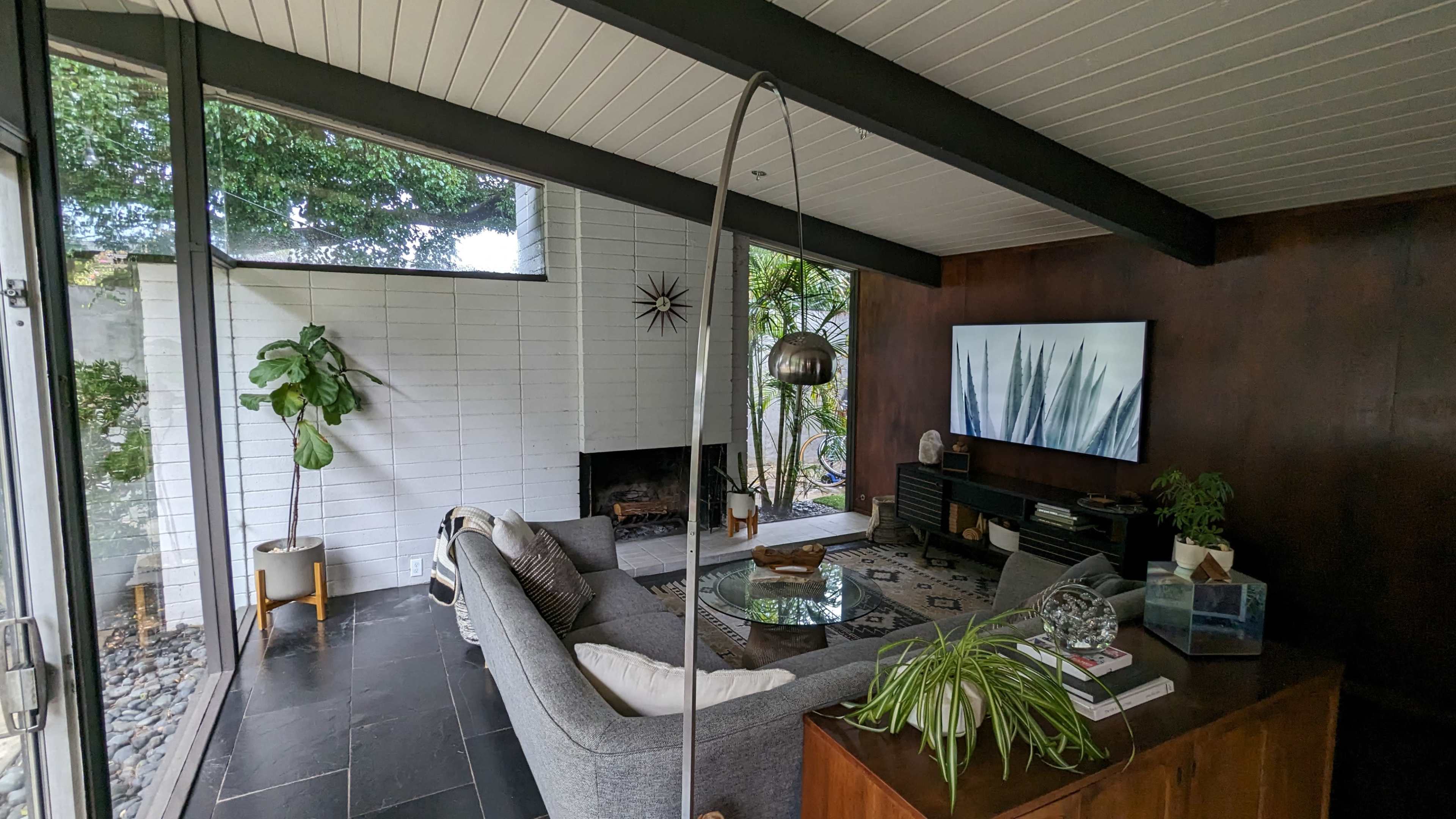 A modern living room features a gray sectional sofa, a glass coffee table, and a large television mounted on the wall, surrounded by large windows and greenery.