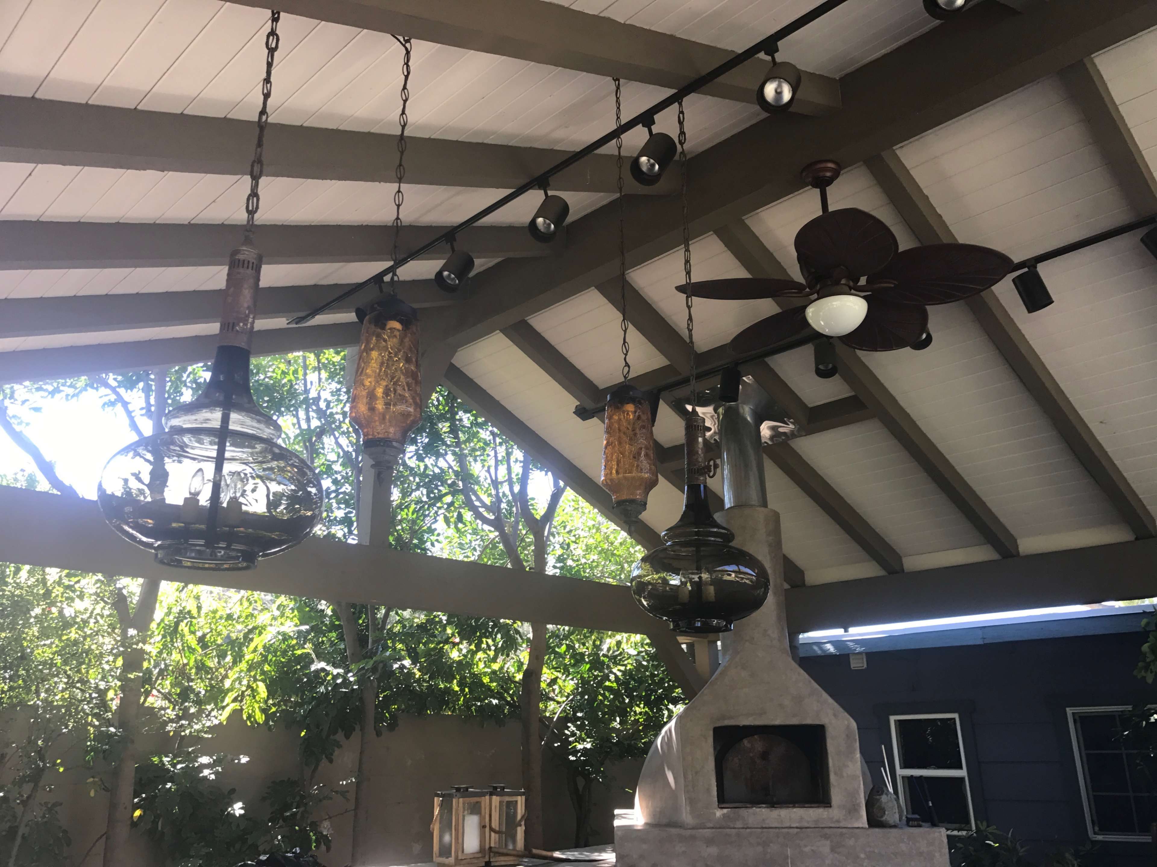 The image shows a covered outdoor area with hanging light fixtures and a ceiling fan, surrounded by greenery.