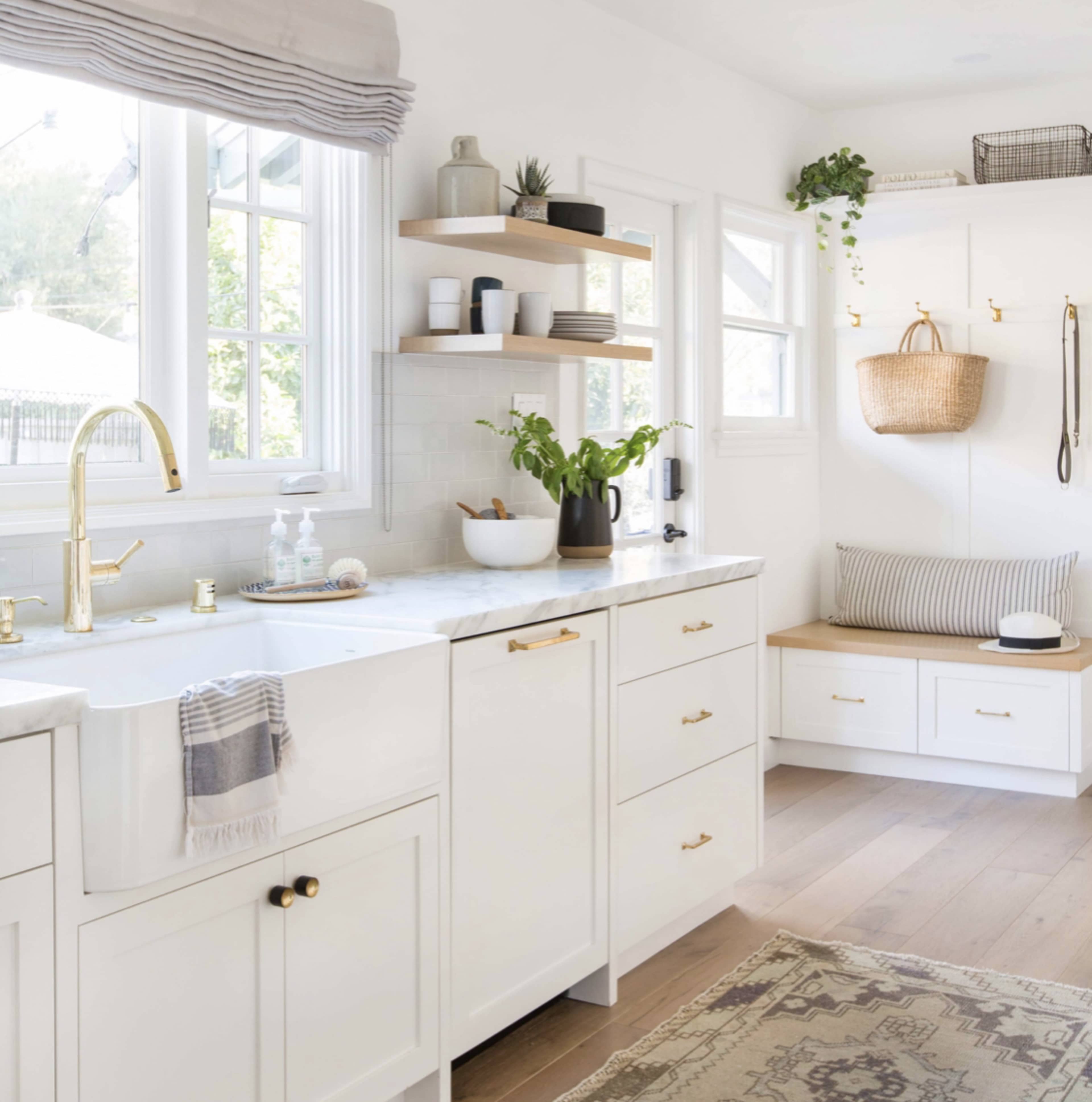 A bright kitchen features white cabinetry, a marble countertop, open shelves with decorative items, and a cozy seating area with a basket and a window.