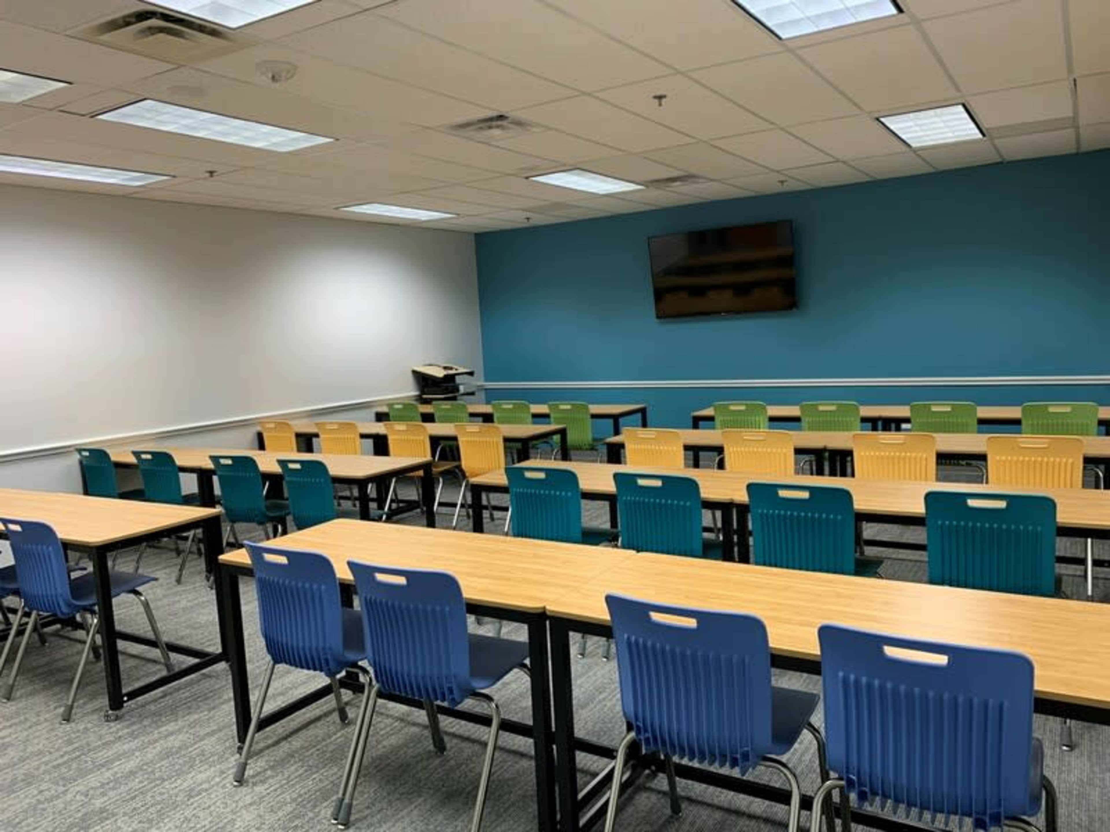 A classroom features several rows of tables with colorful chairs and a wall-mounted television.