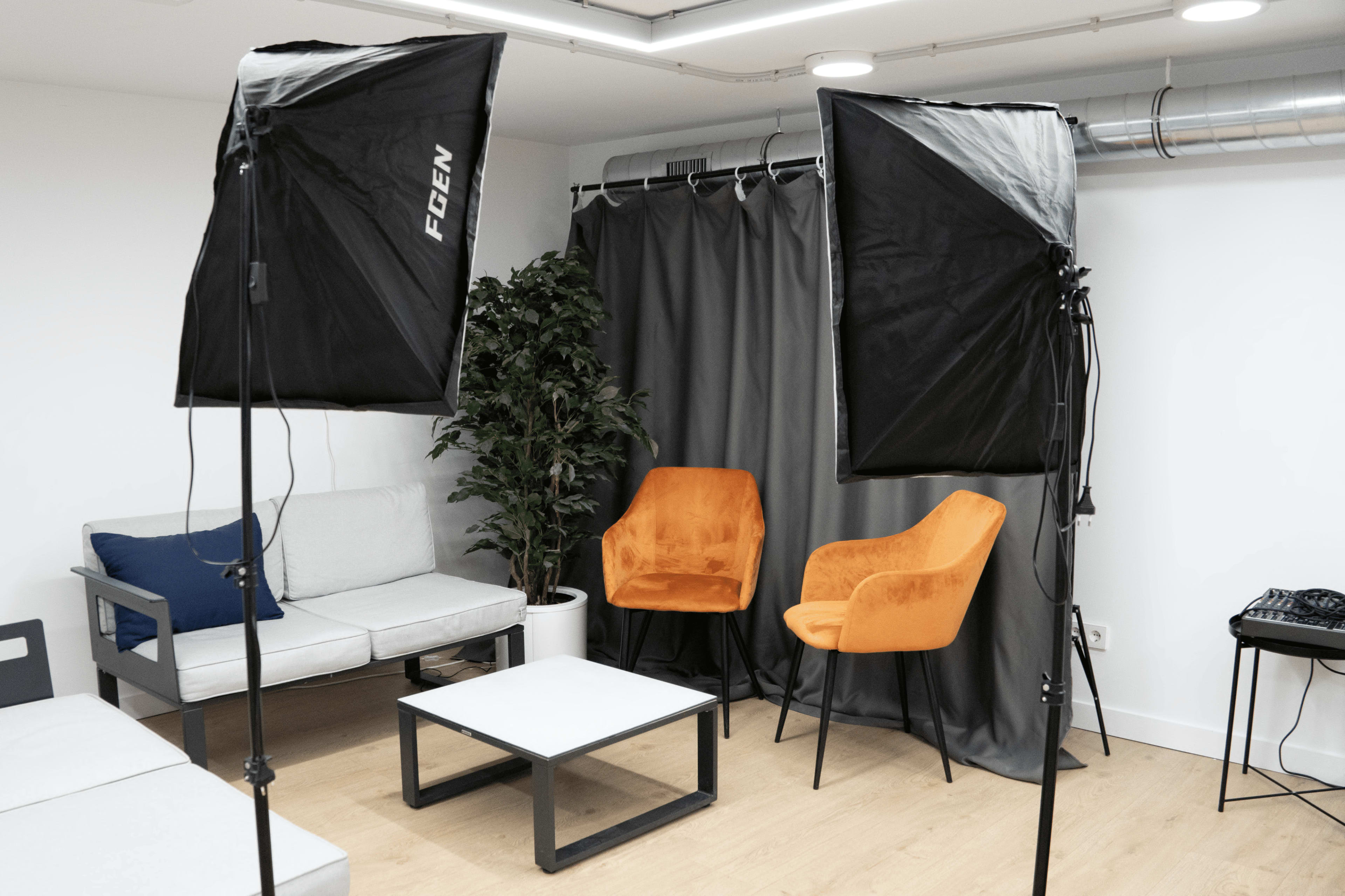 The image shows a minimalist interview setup featuring two orange chairs, a small coffee table, and a gray backdrop, with two softbox lights illuminating the scene.