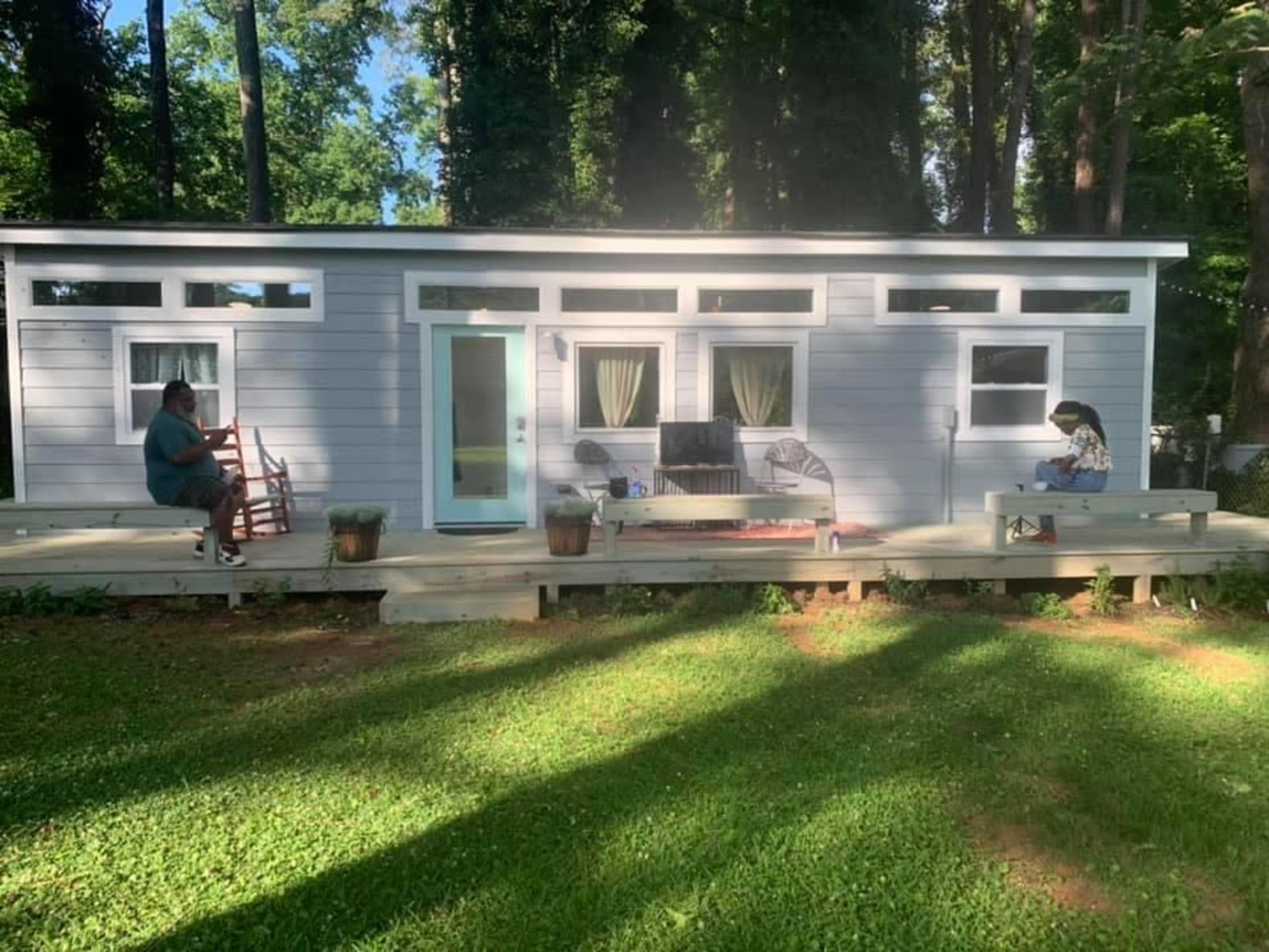 Two people are seated on the deck of a small gray house surrounded by trees.
