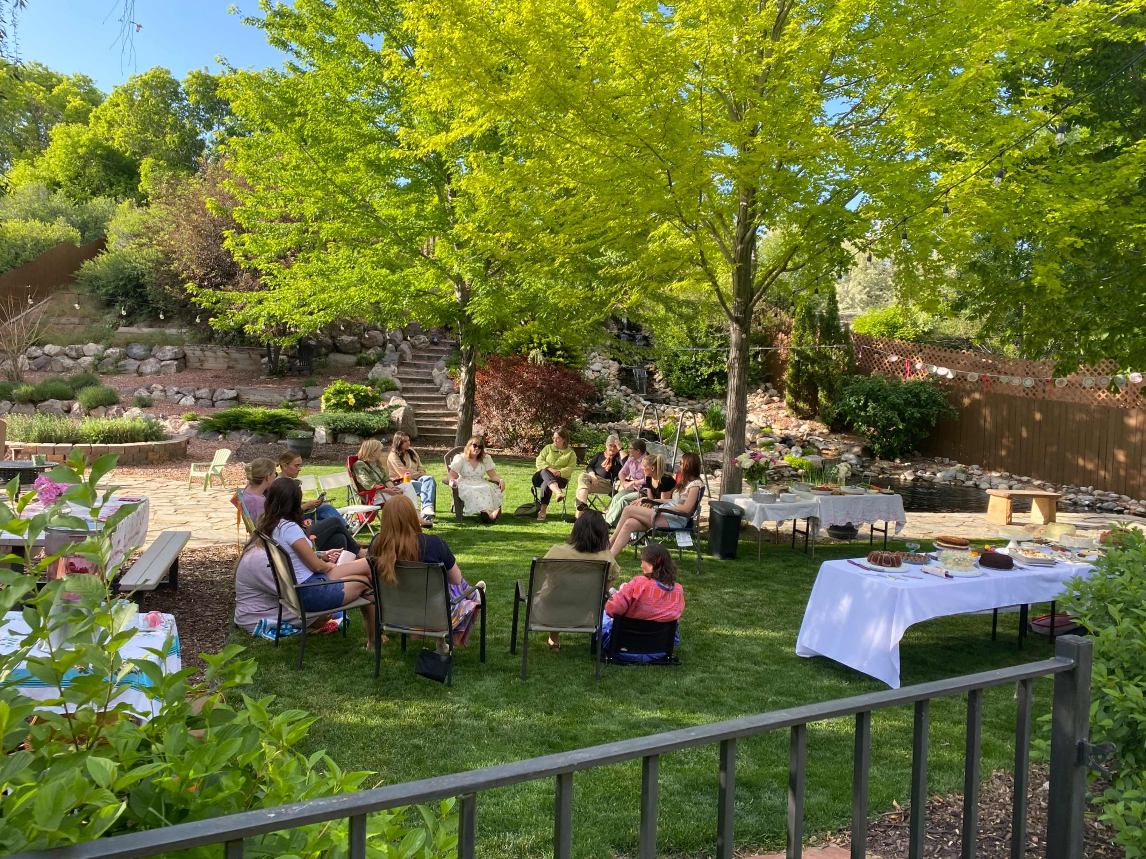 A group of people sits in a circle on a lawn, surrounded by trees and a garden, with tables of food nearby.