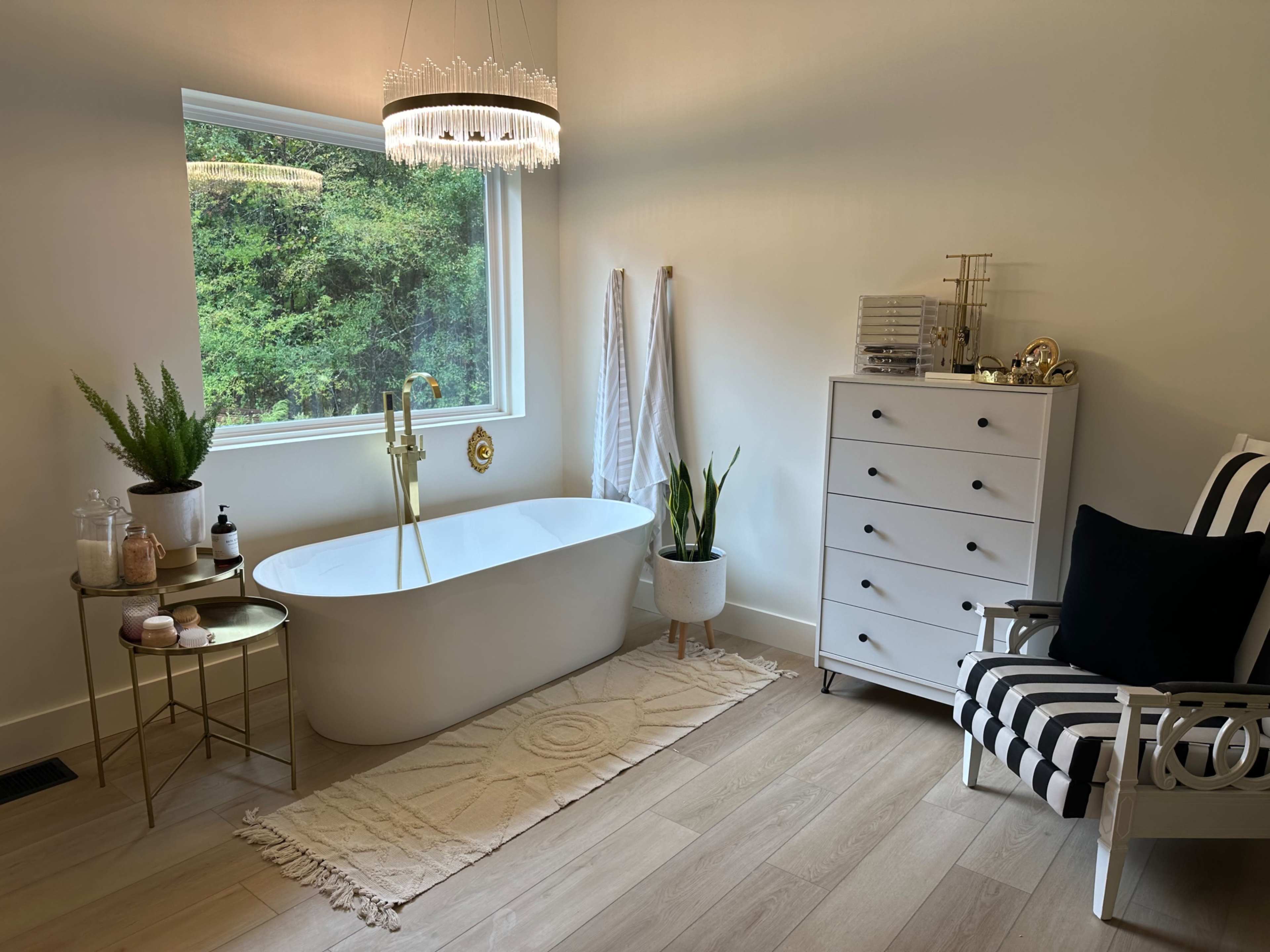 The image shows a modern bathroom featuring a freestanding white bathtub, a large window with greenery outside, a striped chair, a dresser, and decorative plants.