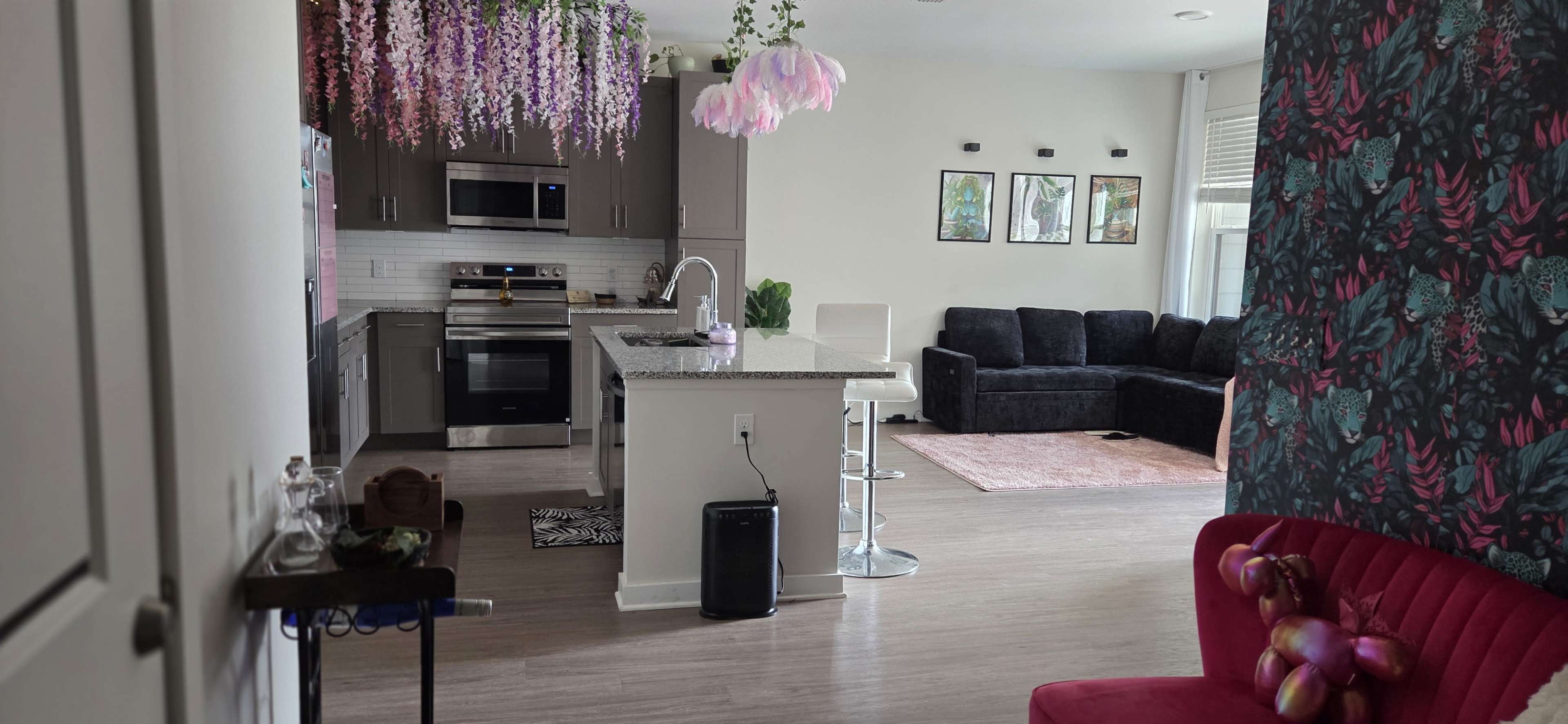 The image shows a modern kitchen with gray cabinetry and stainless steel appliances, adjacent to a living area featuring a black sectional sofa and decorative plants.