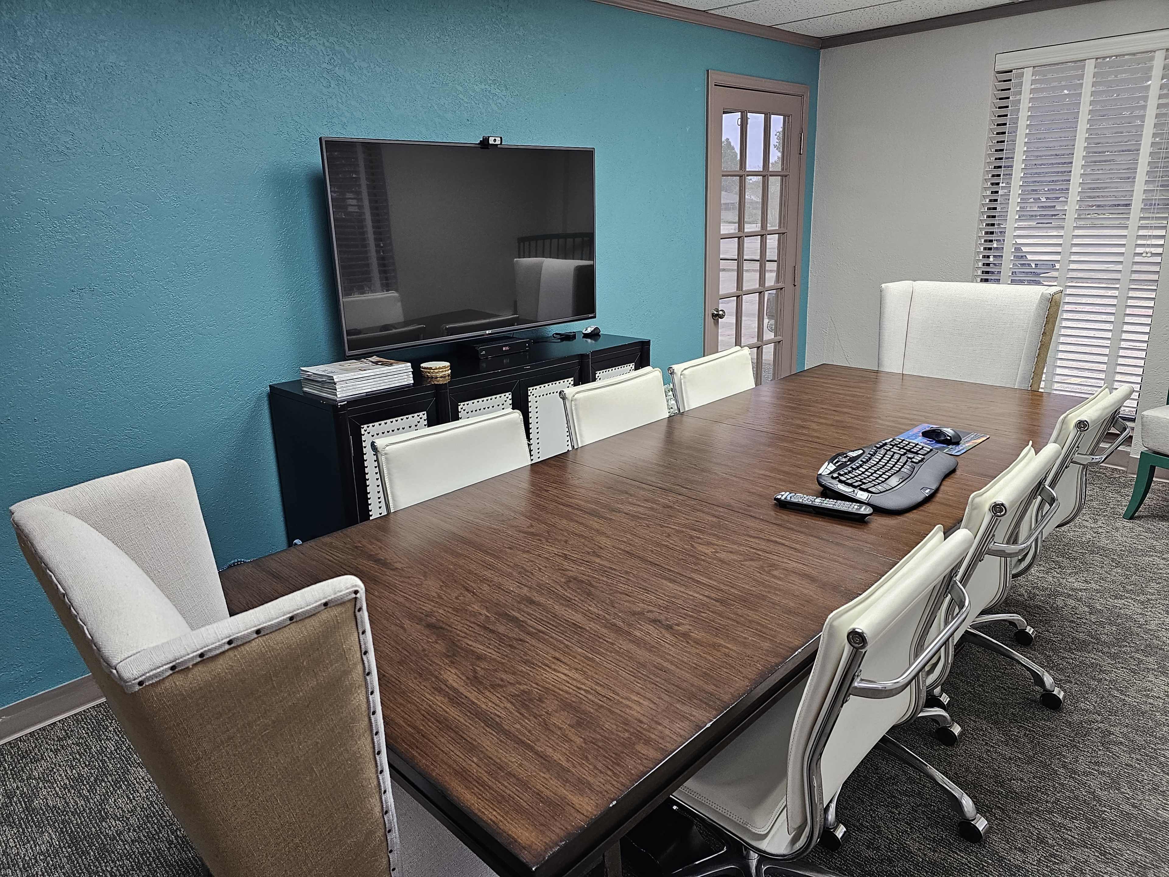 A modern conference room features a long wooden table surrounded by white ergonomic chairs, with a large television mounted on a blue wall.