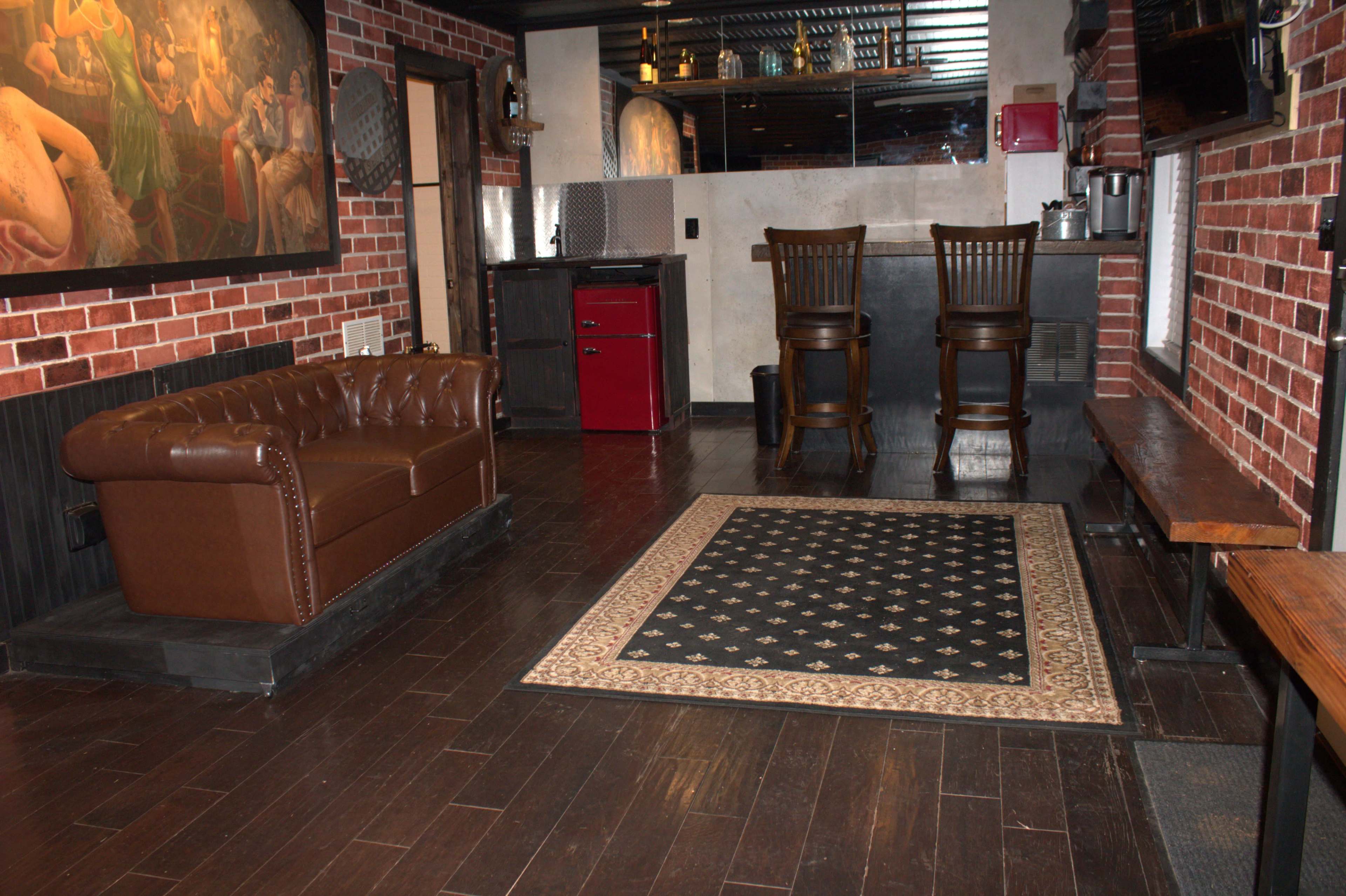 The image shows a small, stylish lounge area featuring a brown leather couch, a patterned rug, a bar with two stools, and exposed brick walls.