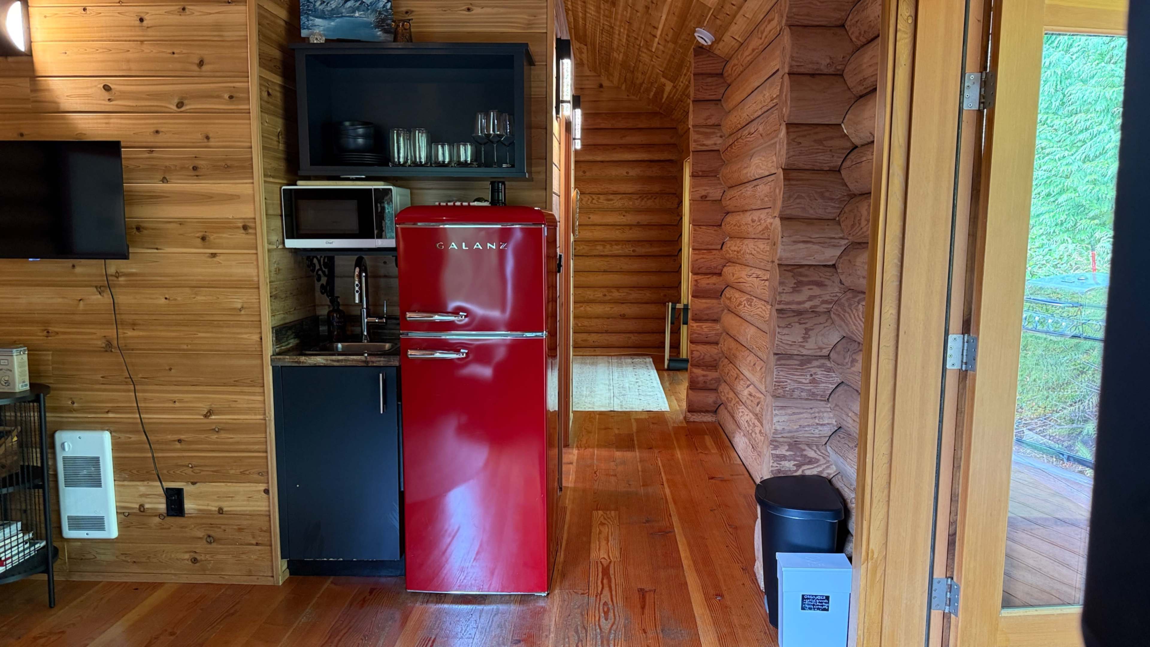 The image shows a wooden interior of a cabin featuring a red refrigerator and a small kitchen area with various kitchen items.