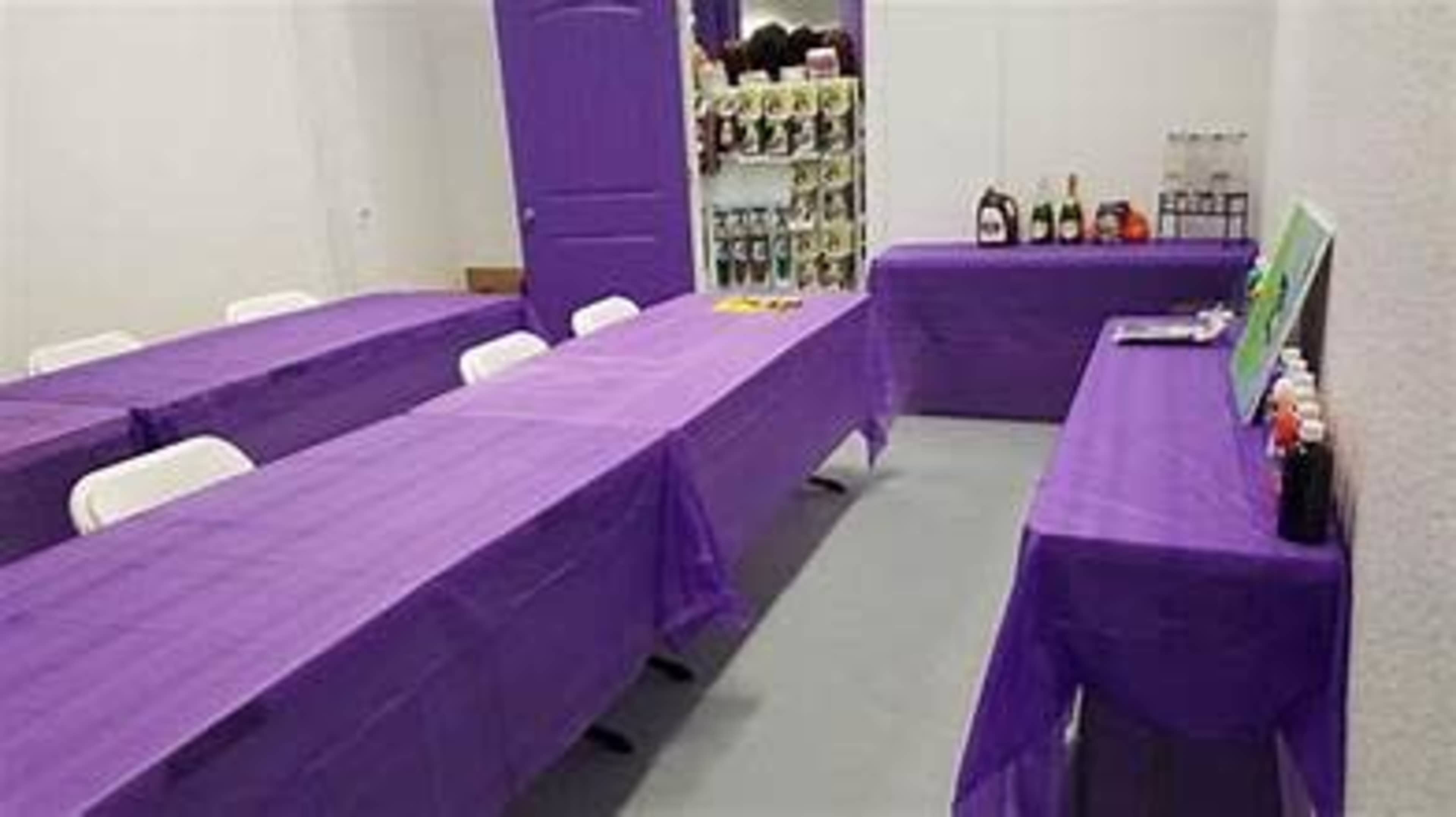 The image shows a room with several tables covered in purple tablecloths, set up for an event, with a shelf of bottles and supplies against one wall.