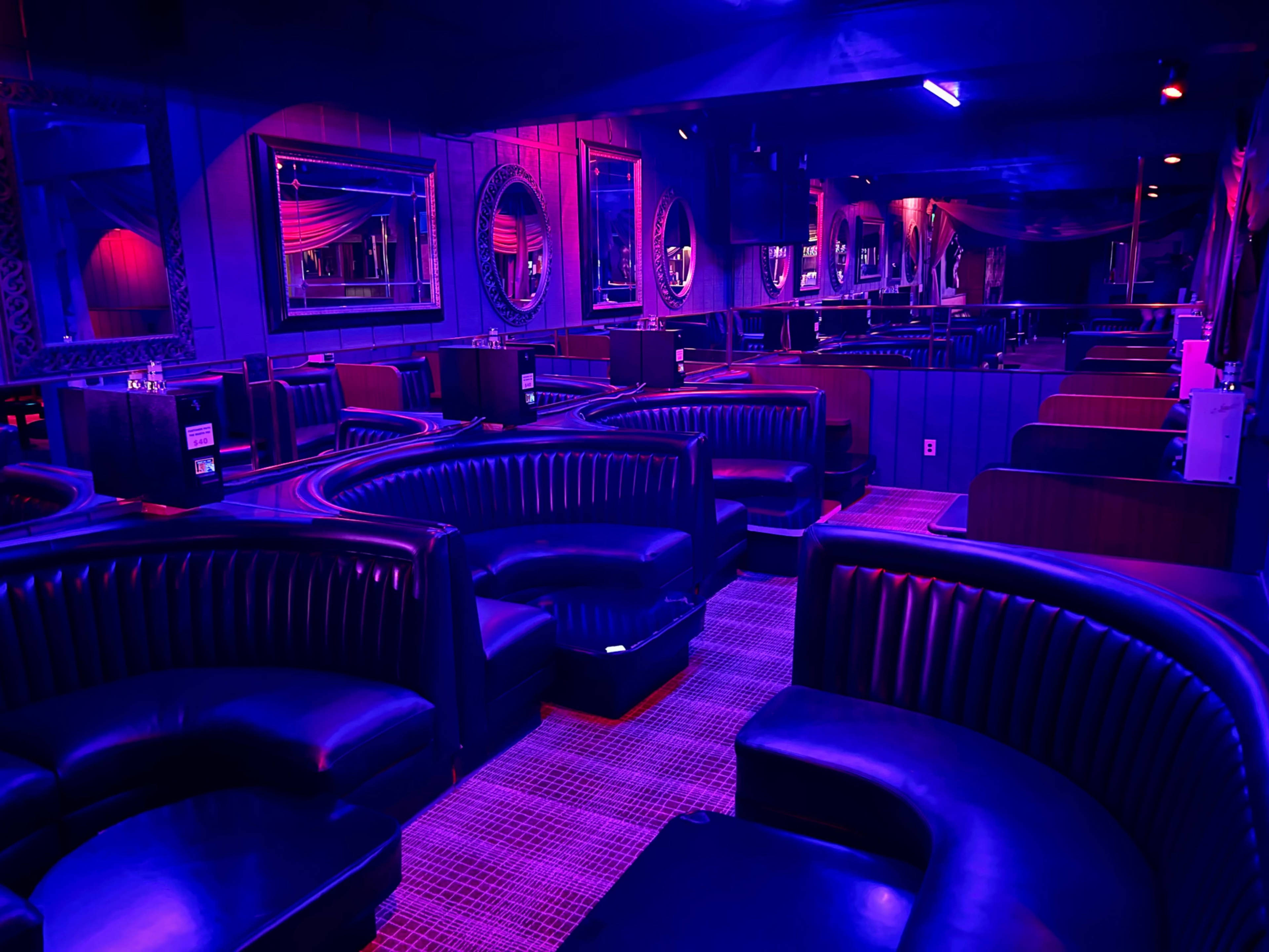An empty lounge with dark leather seating arranged in curved shapes, illuminated by purple and blue lighting.