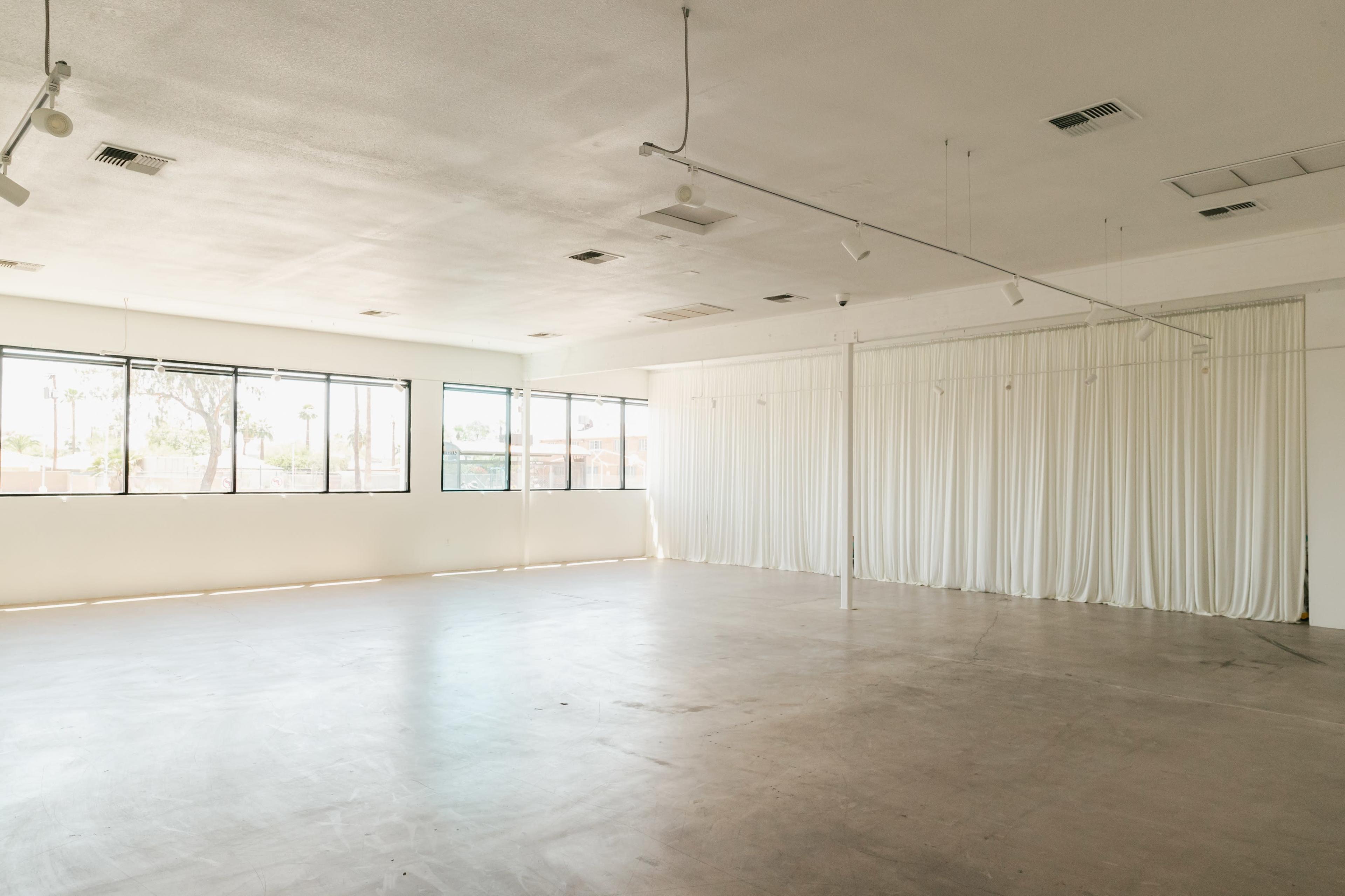The image shows a large, empty room with concrete flooring, large windows on one side, and white curtains along one wall.