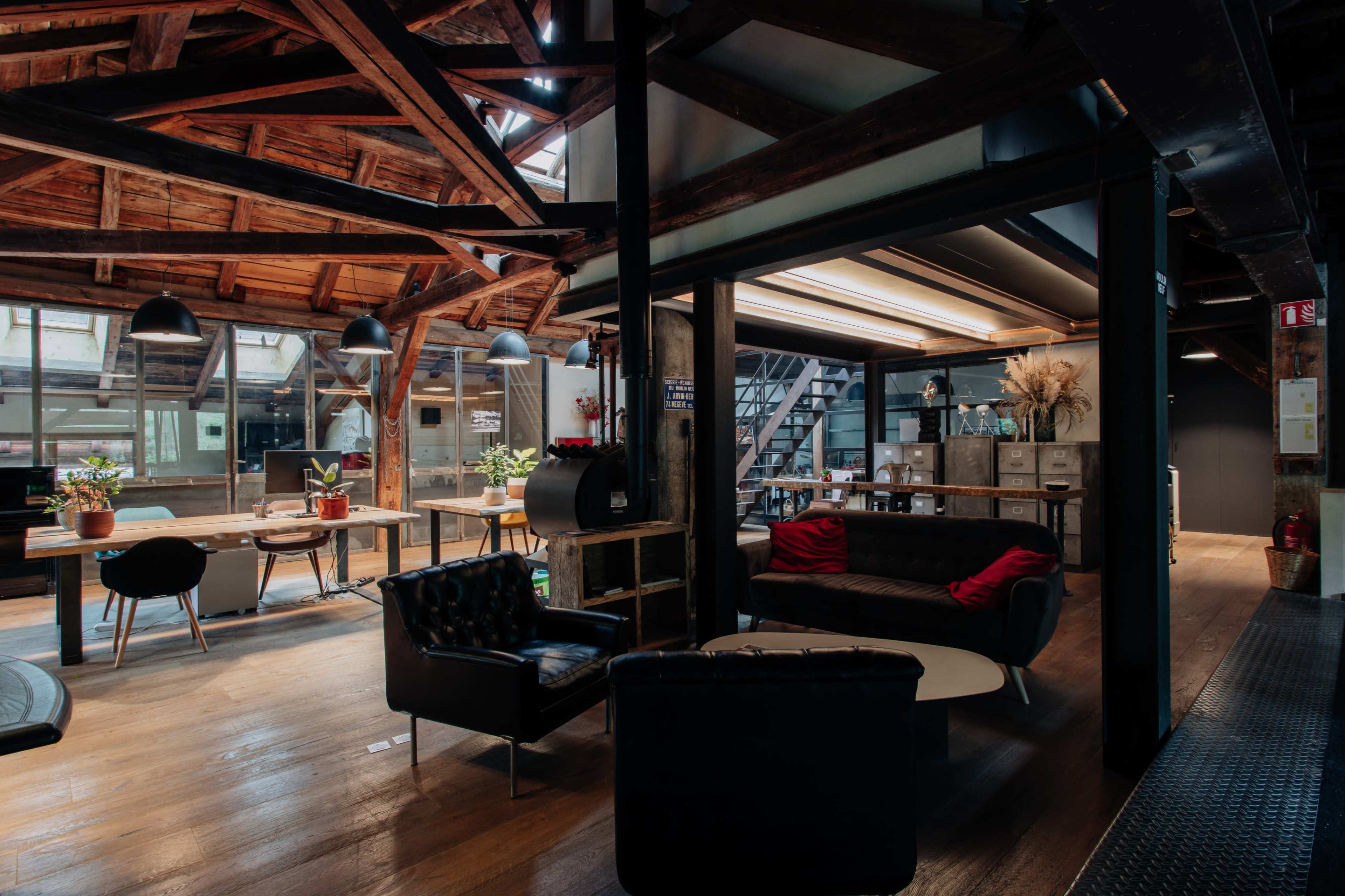 The image shows a modern open-plan workspace with wooden beams and a mix of seating areas, including tables, chairs, and couches.