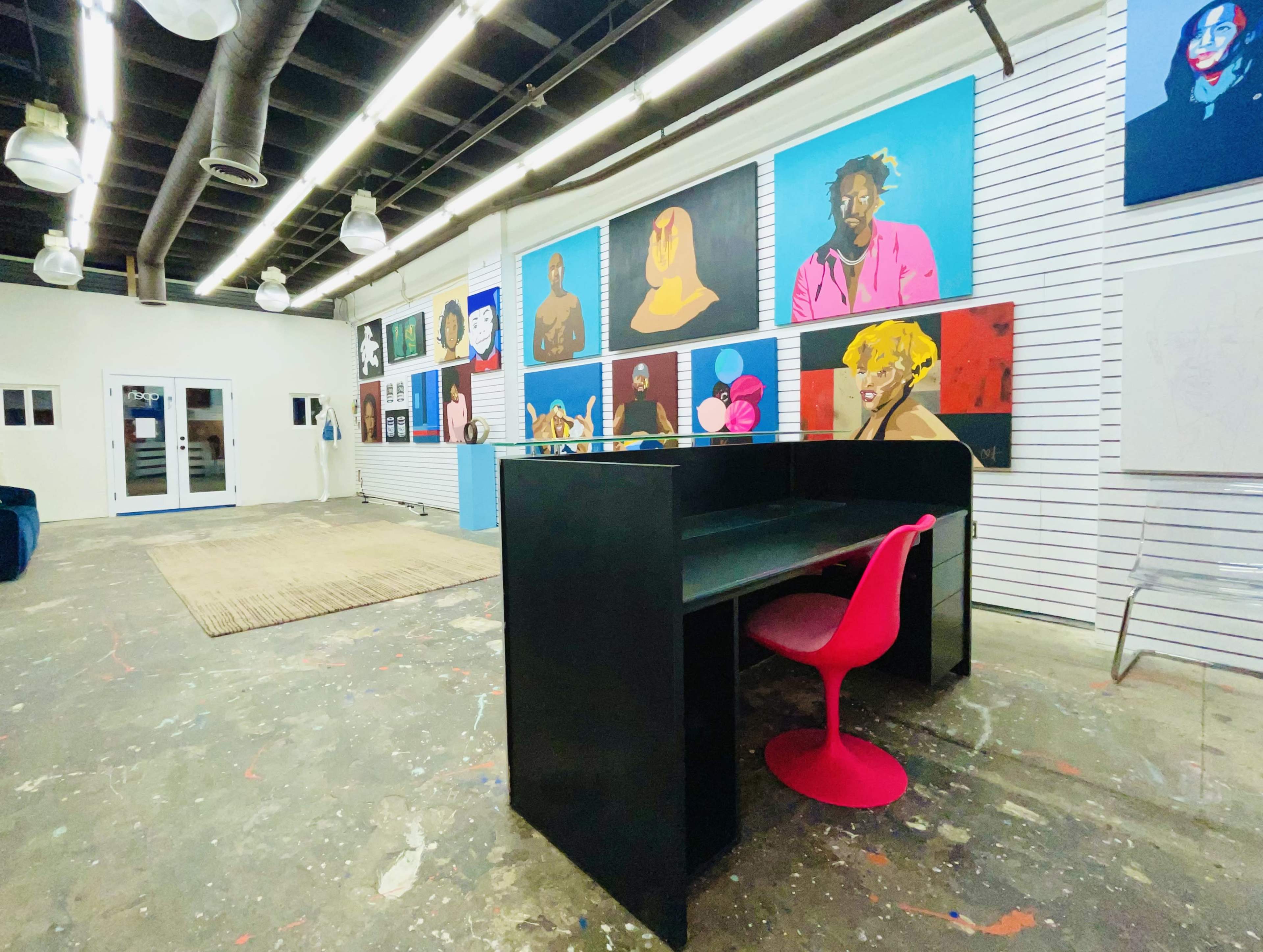 The image shows a spacious gallery-like room with colorful artwork displayed on the walls, featuring a black desk and a pink chair in the foreground.