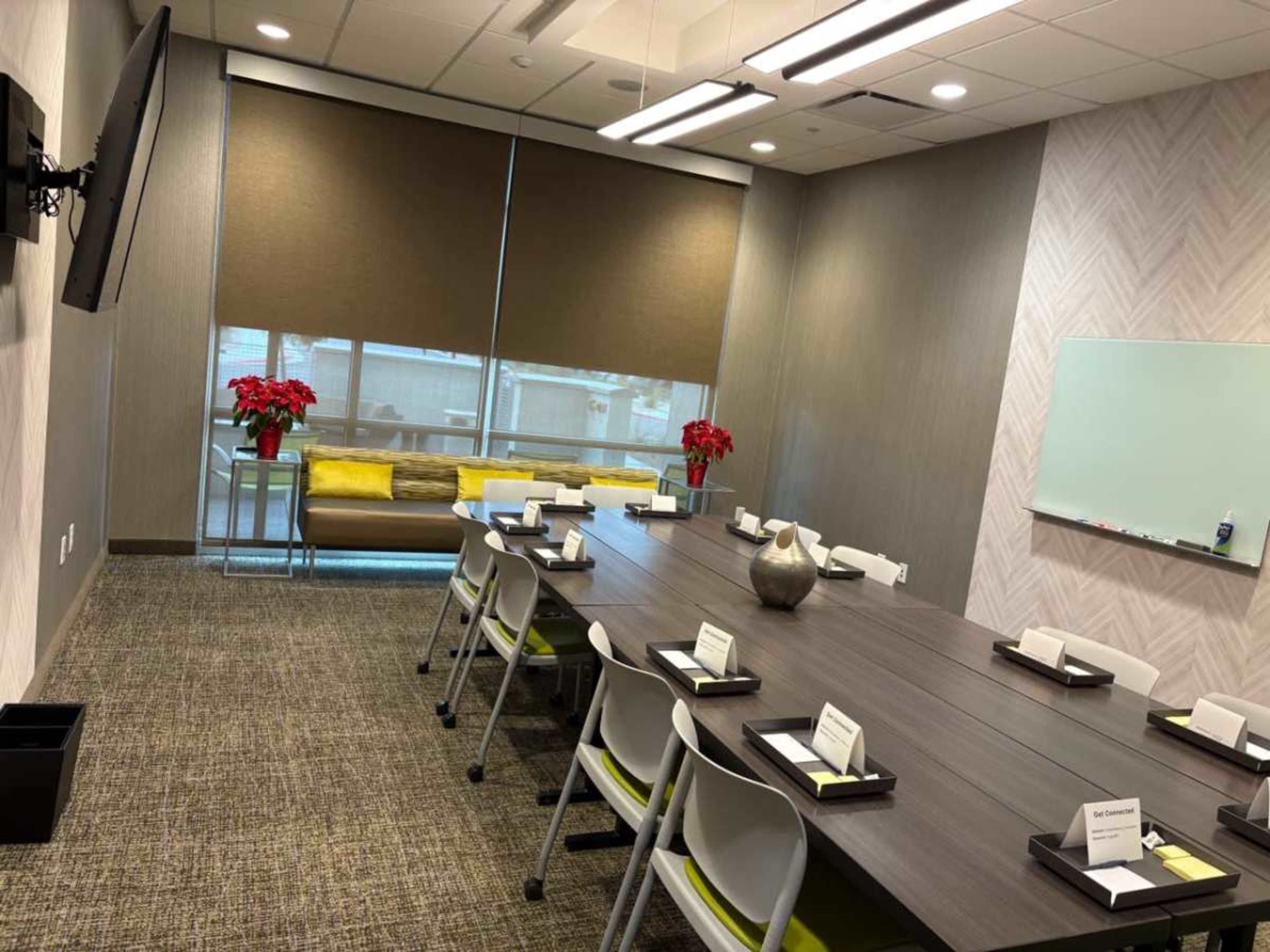 A modern conference room features a long table with chairs, a television on the wall, and large windows with shades, adorned with potted plants.