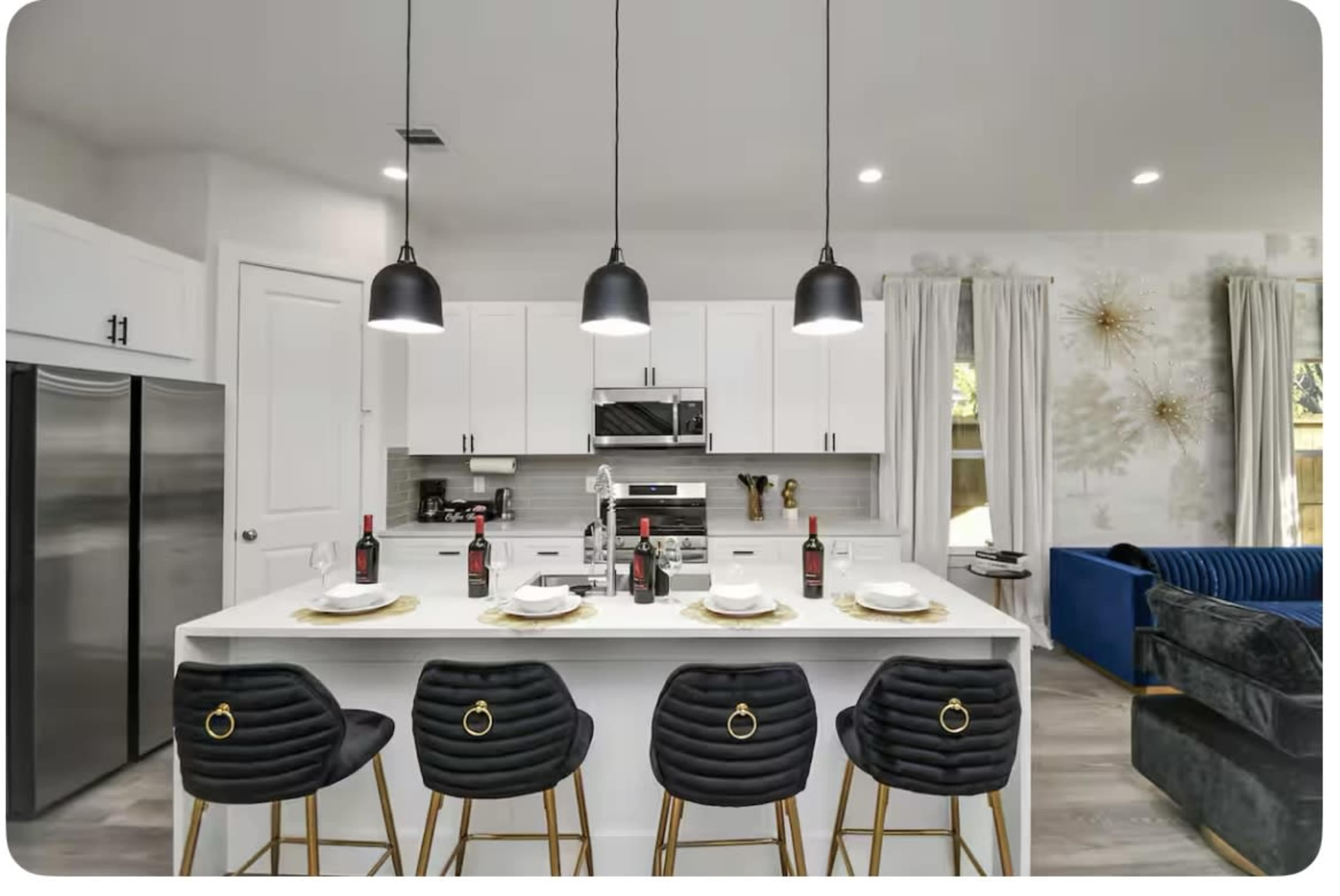 A modern kitchen features a center island with high stools and pendant lighting, equipped with stainless steel appliances and a dining setup.