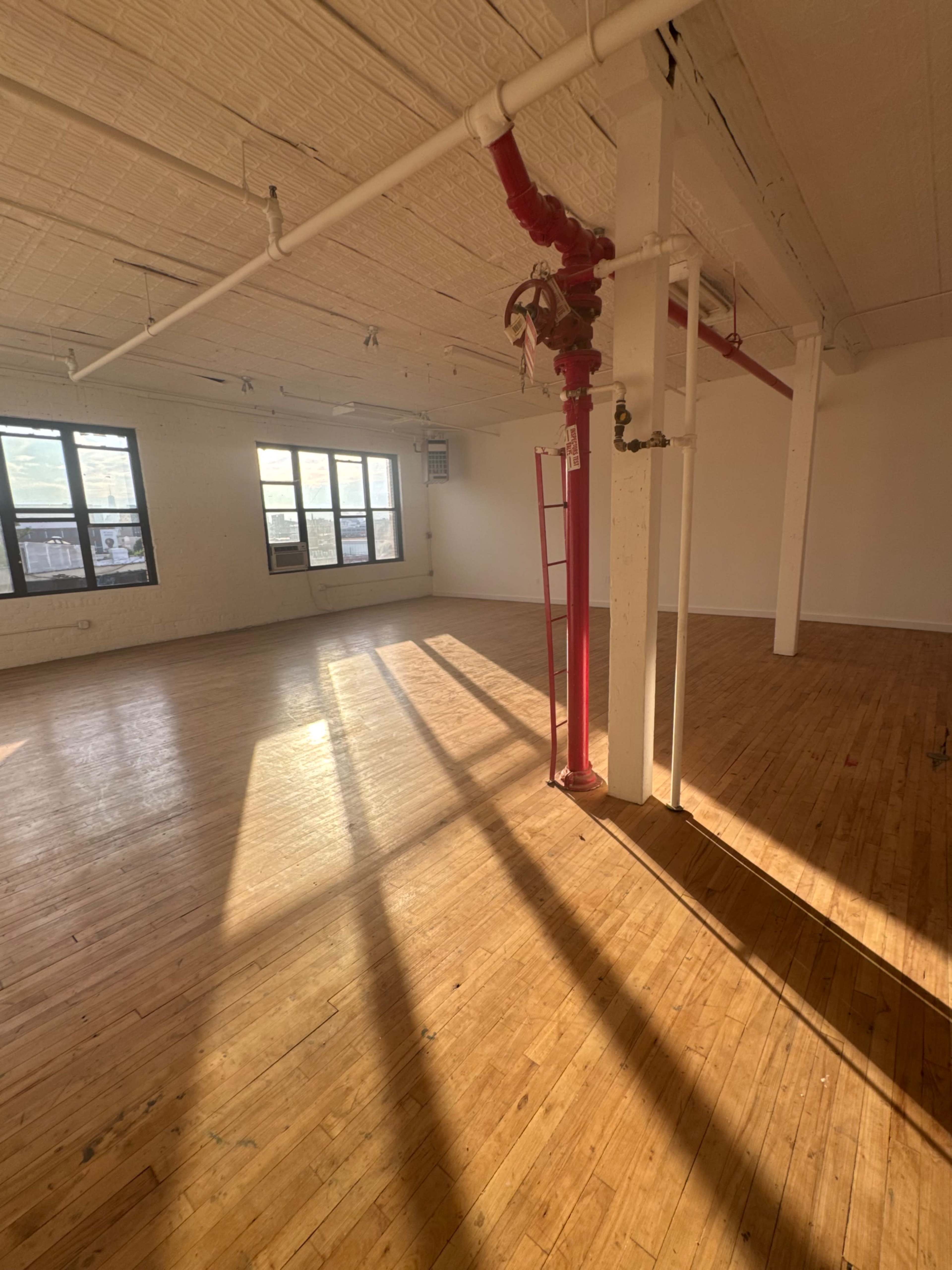 The image shows a large, empty room with wooden floors, large windows allowing sunlight to enter, and exposed pipes along the walls.
