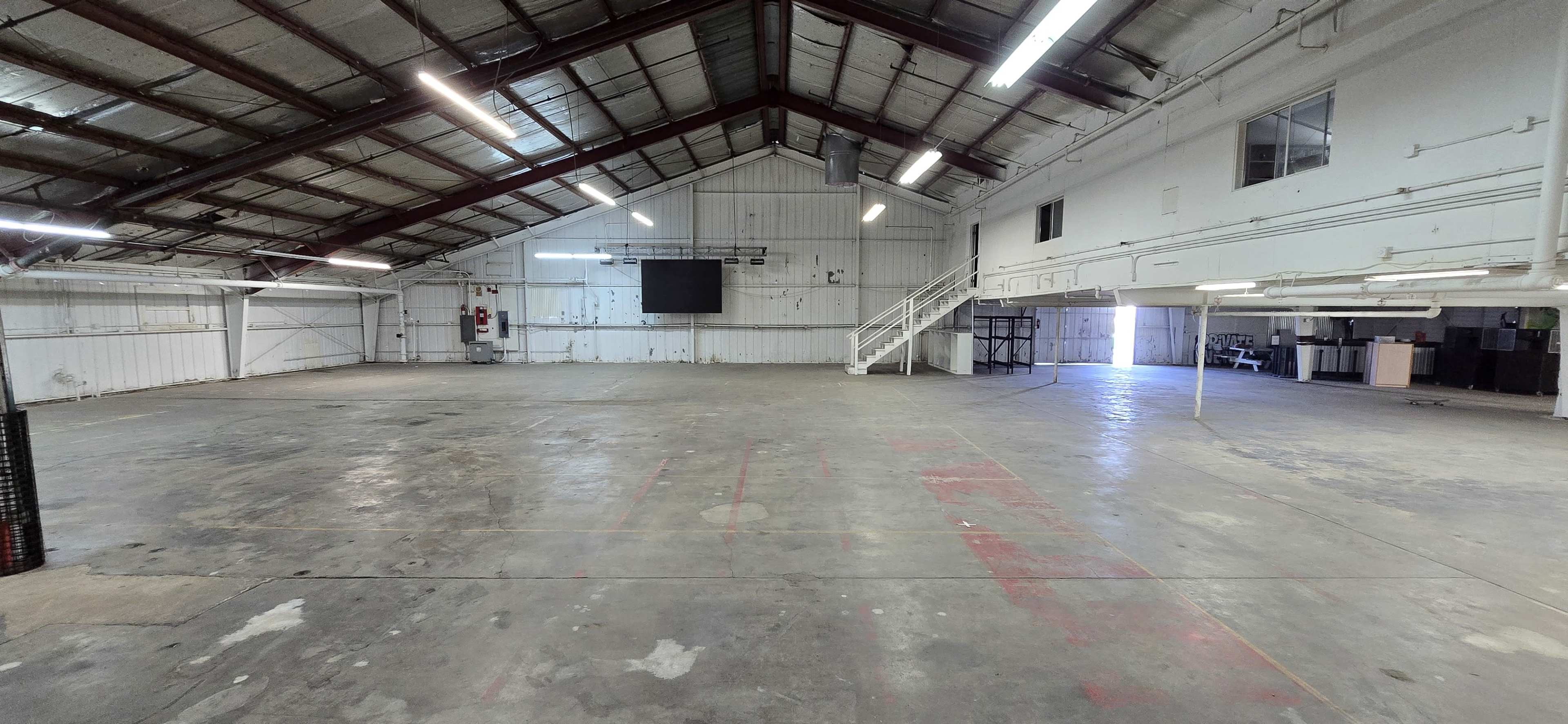 The image shows a large, empty industrial space with a high ceiling, featuring exposed beams and minimal furnishings.