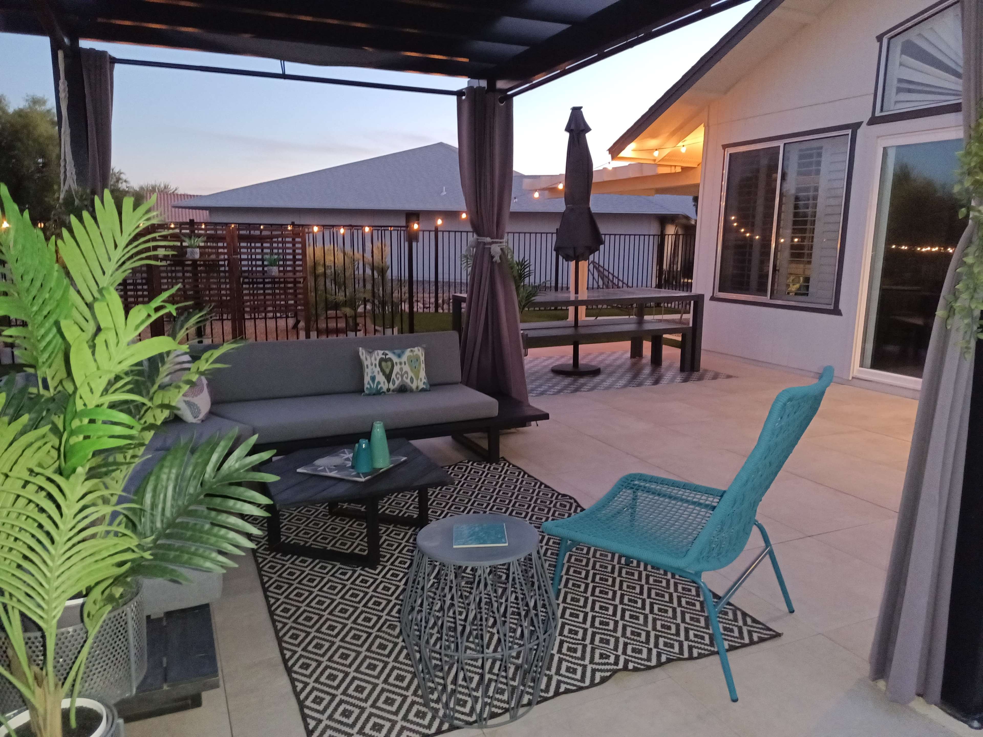 A modern outdoor patio area with a gray sofa, a patterned rug, a blue chair, and potted plants, set against a backdrop of a house and twilight sky.