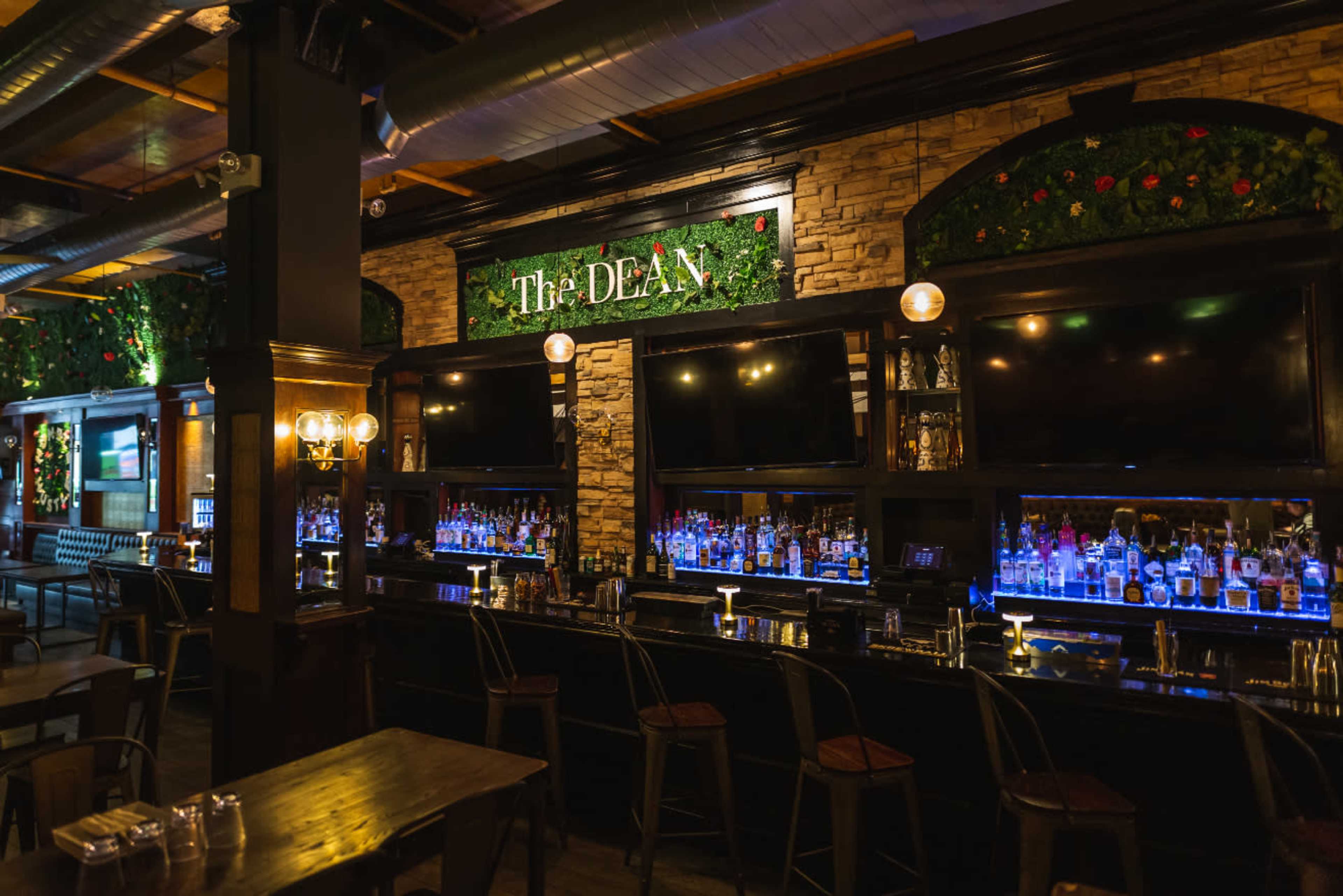 The image shows a bar area featuring a dark wood counter with several bar stools and shelves lined with liquor bottles, illuminated by ambient lighting.