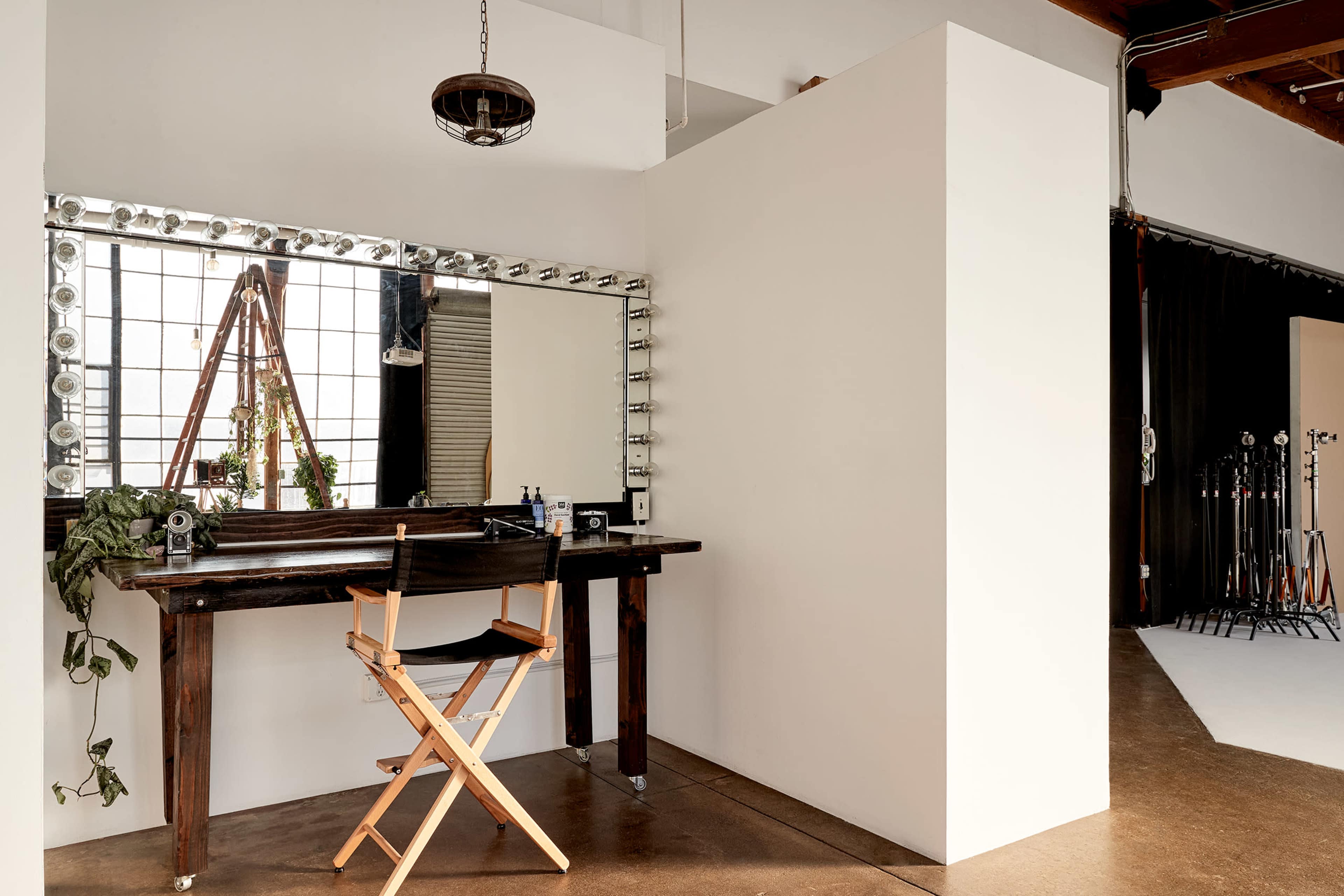 The image shows a minimalist makeup station featuring a large mirror with lights, a wooden chair, and a white partition in a well-lit room.