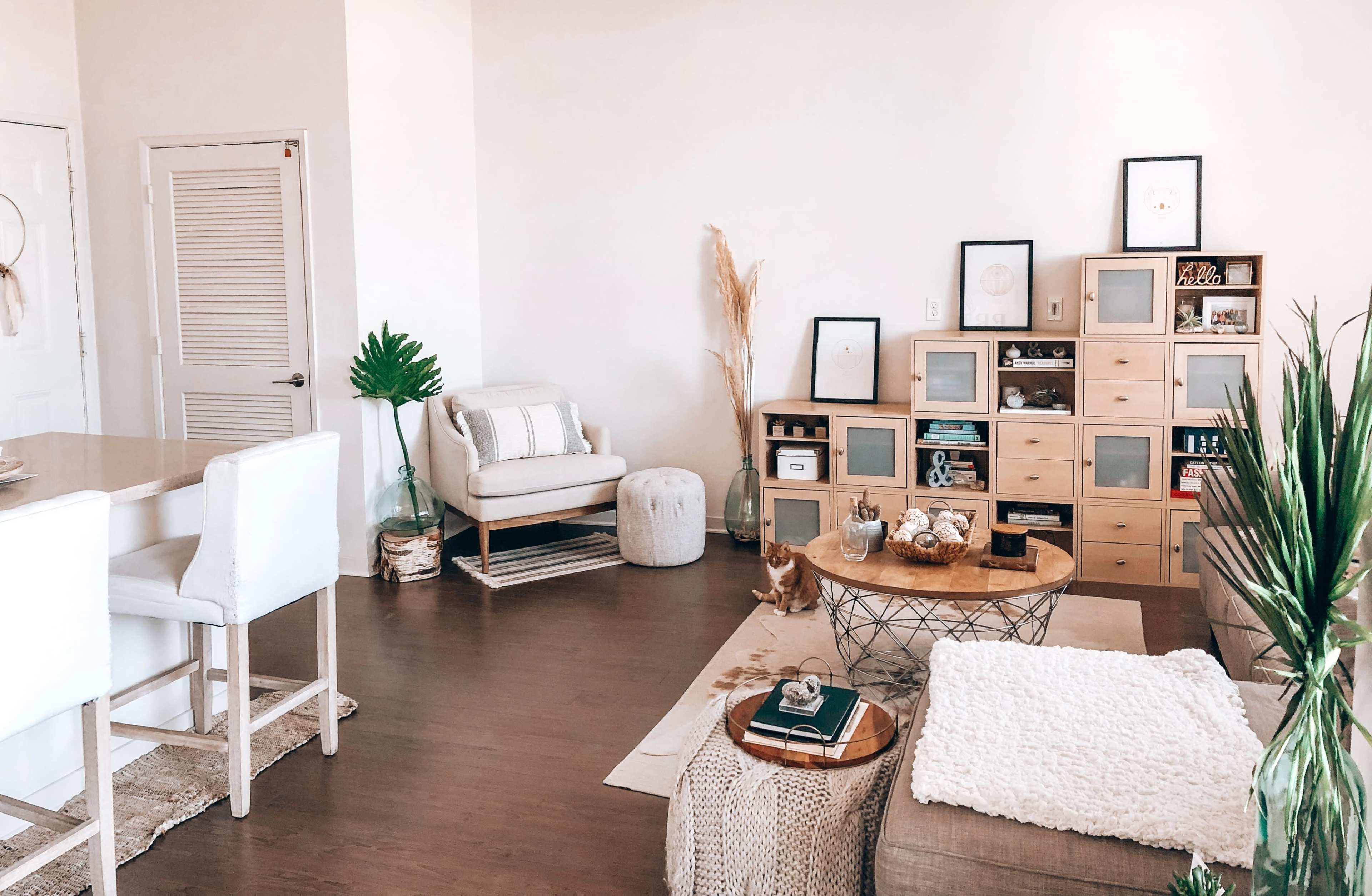 A cozy living space features a light-colored armchair, a wooden bookshelf with decorative items, a round coffee table, and a small dining area.