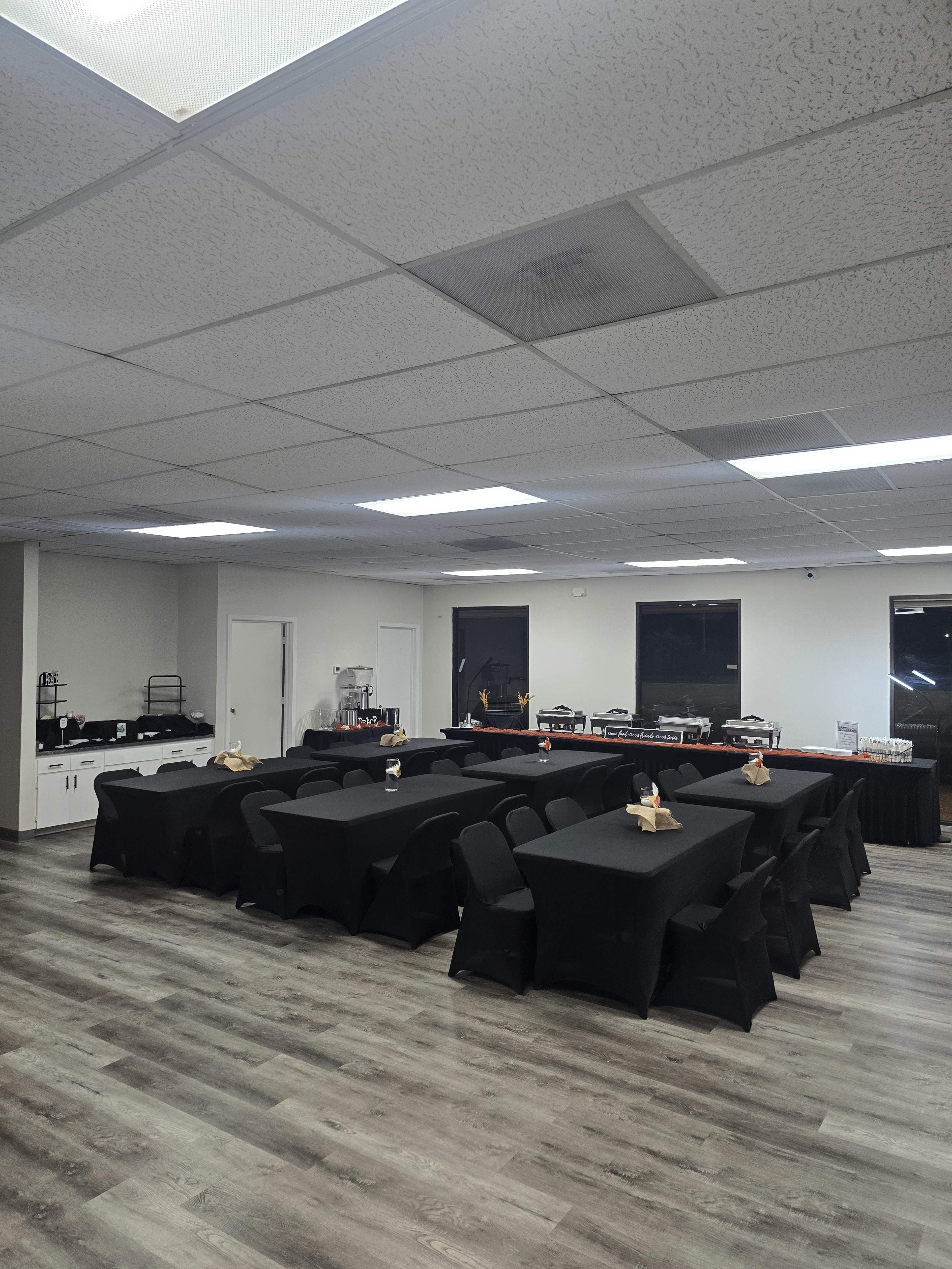 The image shows a spacious indoor area set up with multiple black-covered tables arranged in a U-shape, surrounded by large windows.