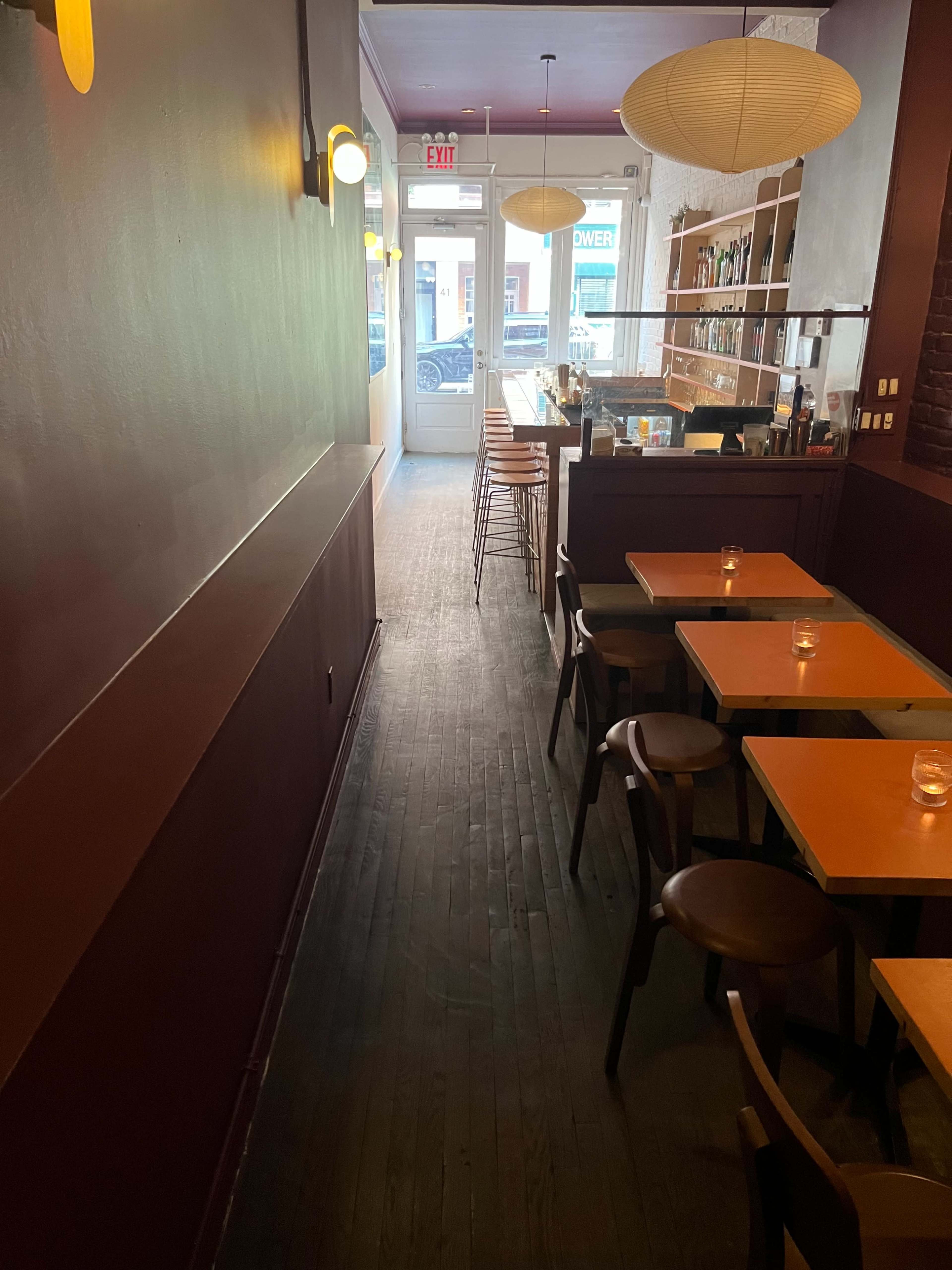 The image shows the interior of a restaurant featuring wooden flooring, empty tables with chairs, and a bar area illuminated by overhead lighting.