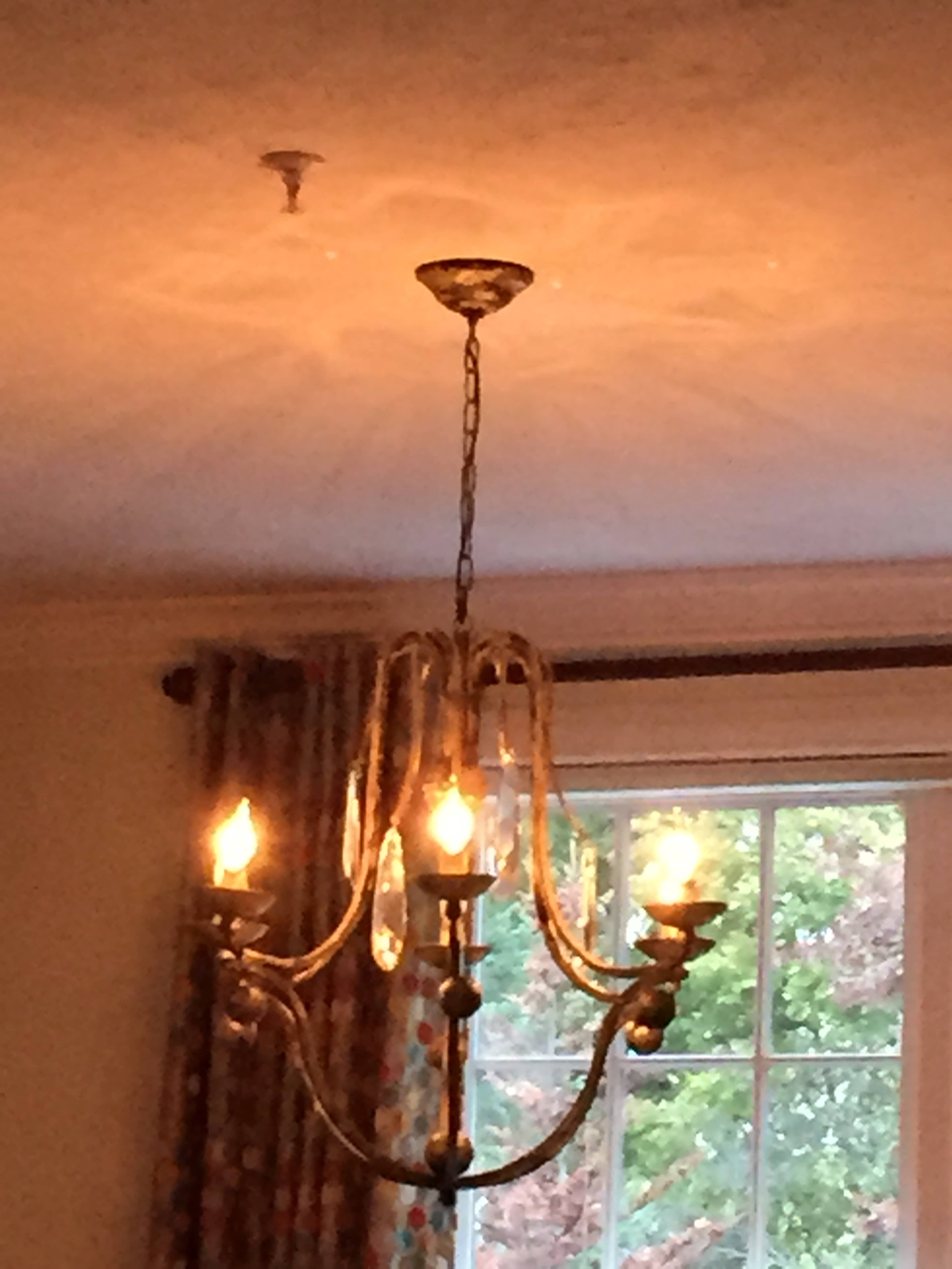 A chandelier with glowing bulbs hangs from the ceiling, casting light patterns on the wall in a room with a window and patterned curtains.