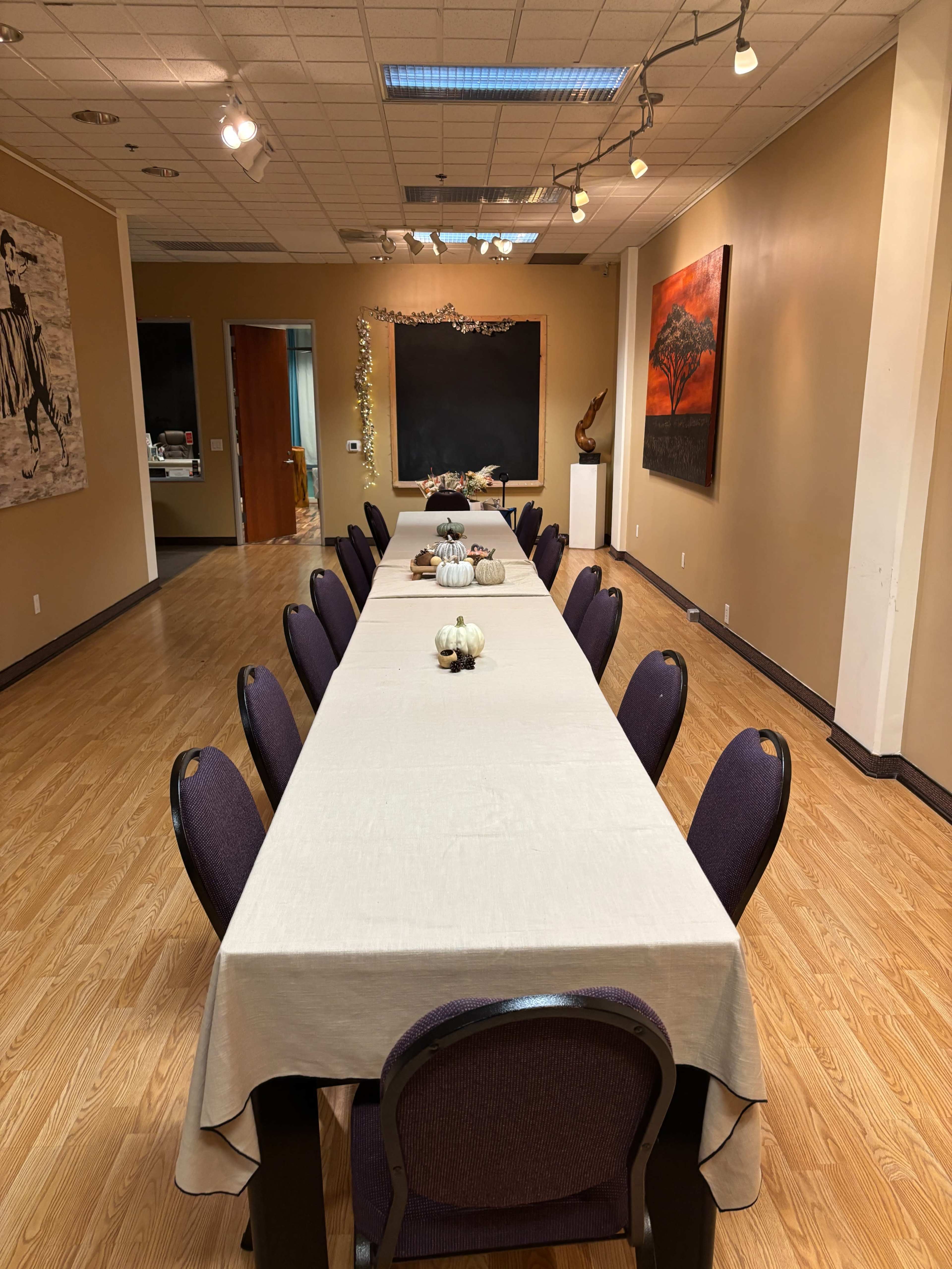 A long banquet table is set with a beige tablecloth and decorative items, surrounded by chairs in a well-lit room with artwork on the walls.