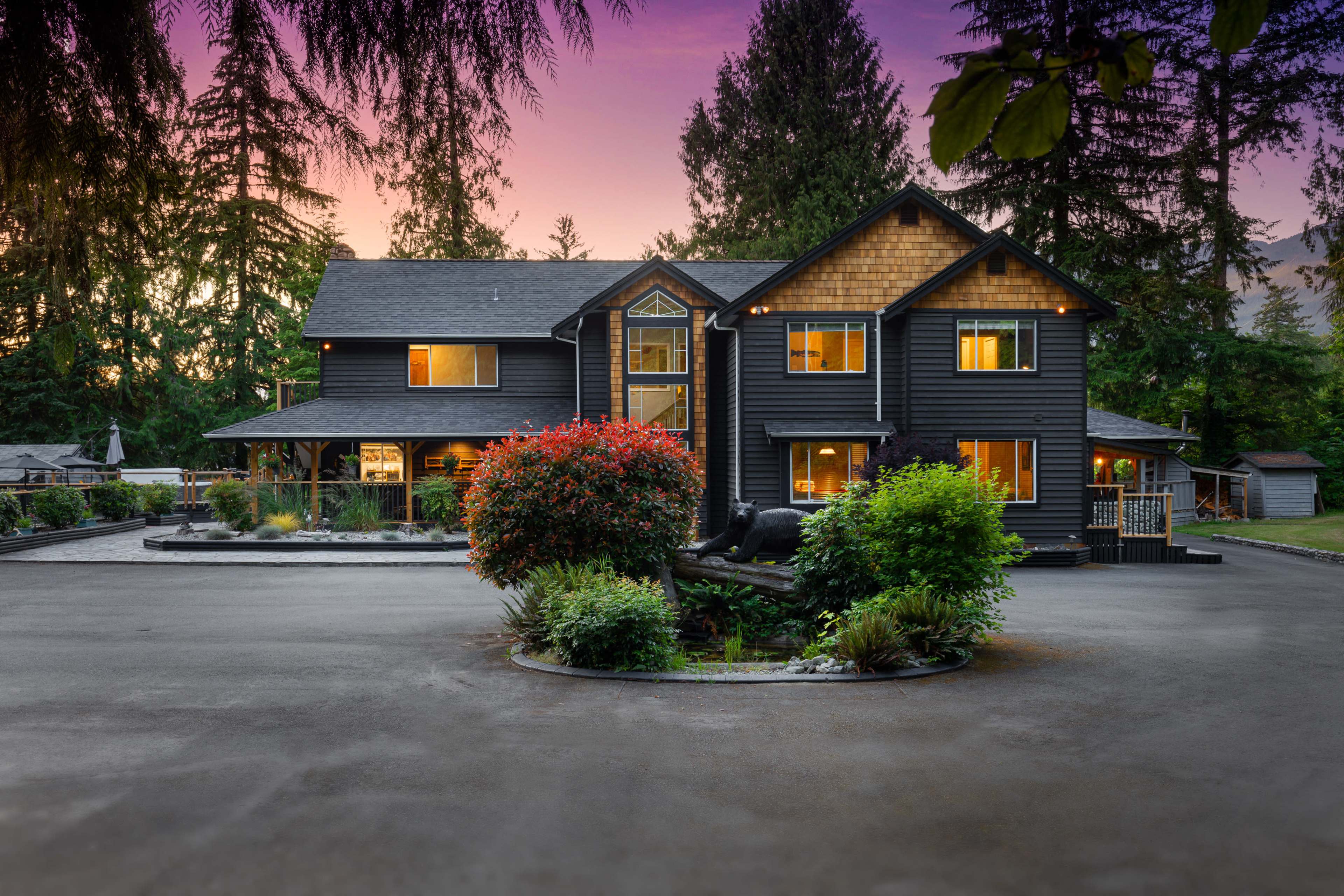 A large black house with multiple windows and a landscaped front garden is set against a backdrop of tall trees at dusk.