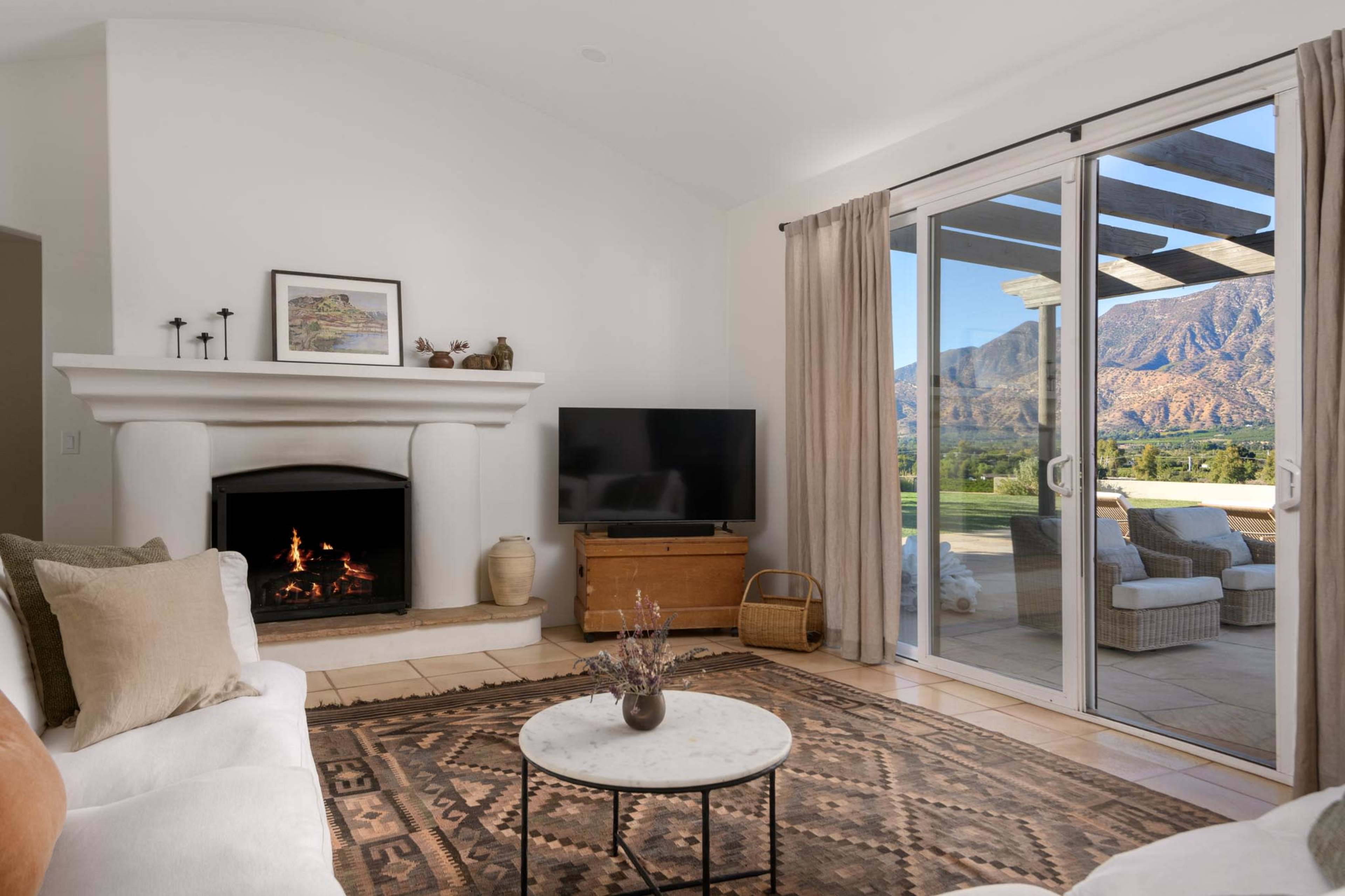 The living room features a white sofa, a round marble coffee table, a television, and a fireplace, with large sliding glass doors providing a view of mountains and outdoor seating.
