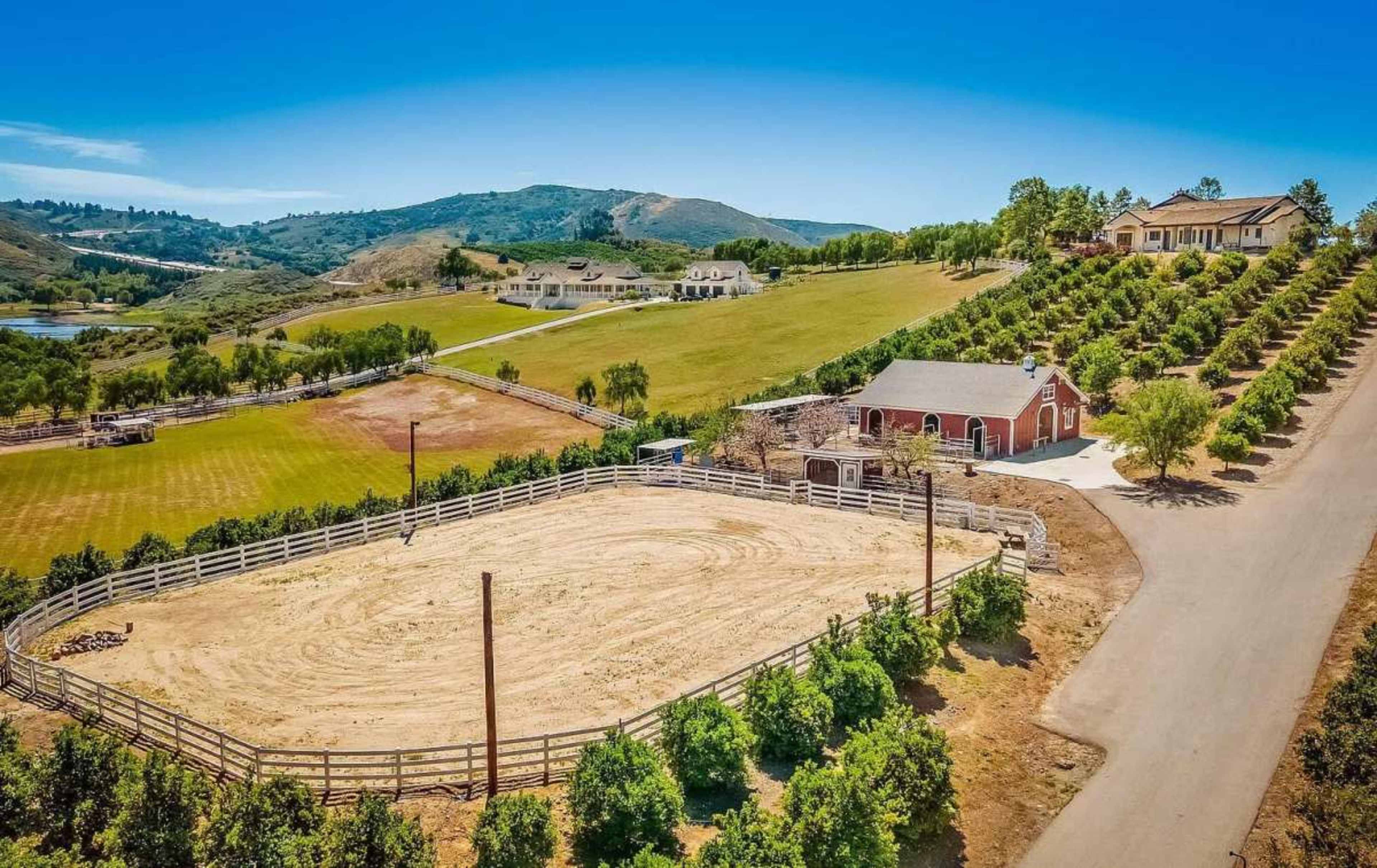 The image shows a spacious rural landscape featuring a red barn, a sandy riding area, and rows of trees on a hillside.