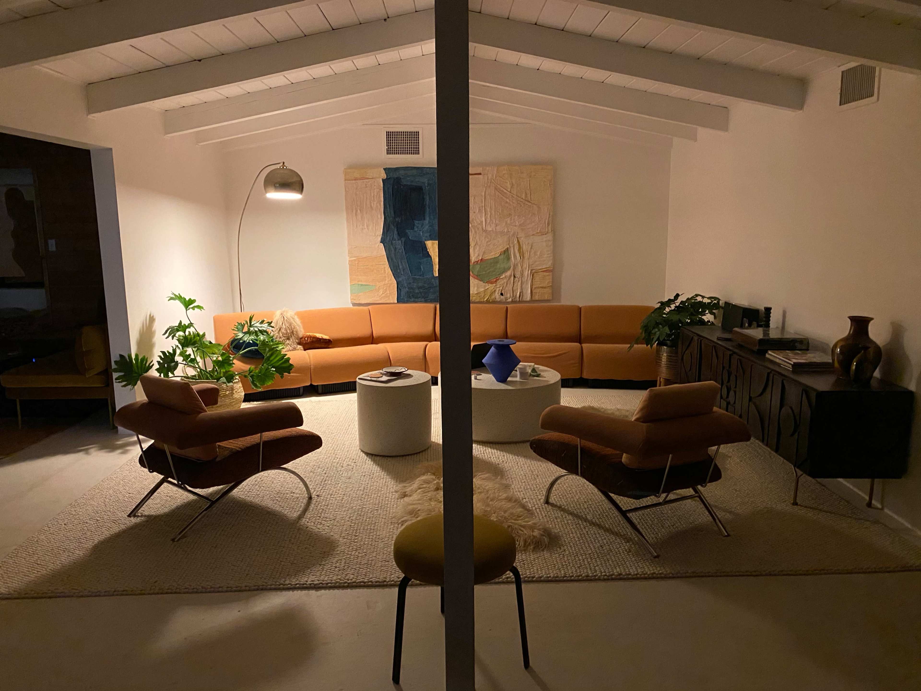 A modern living room with a curved orange sofa, two brown armchairs, a round white coffee table, and various plants, all under a ceiling with wooden beams.