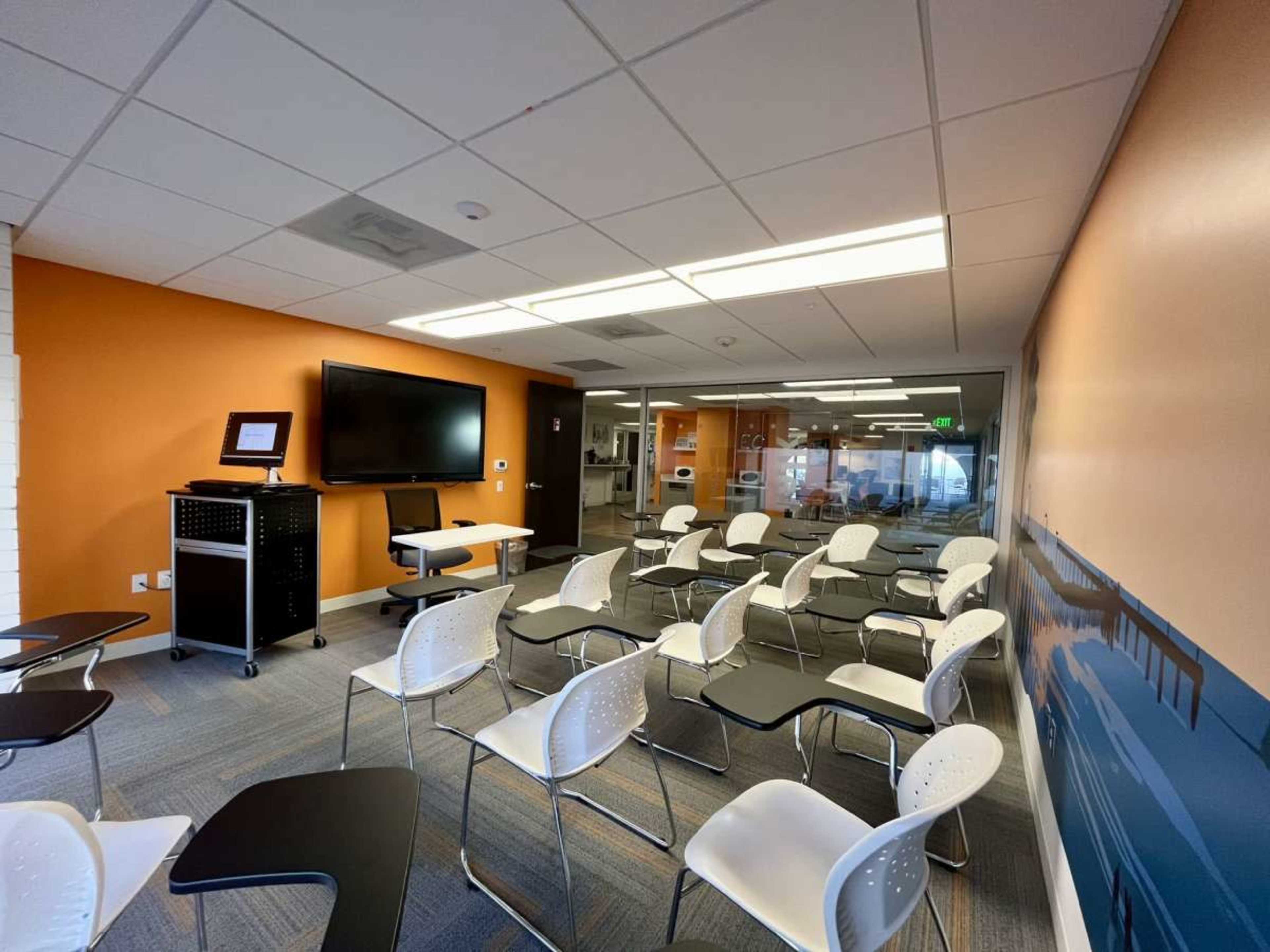 A modern classroom features multiple white chairs arranged in rows, with a blackboard and a television mounted on the wall.