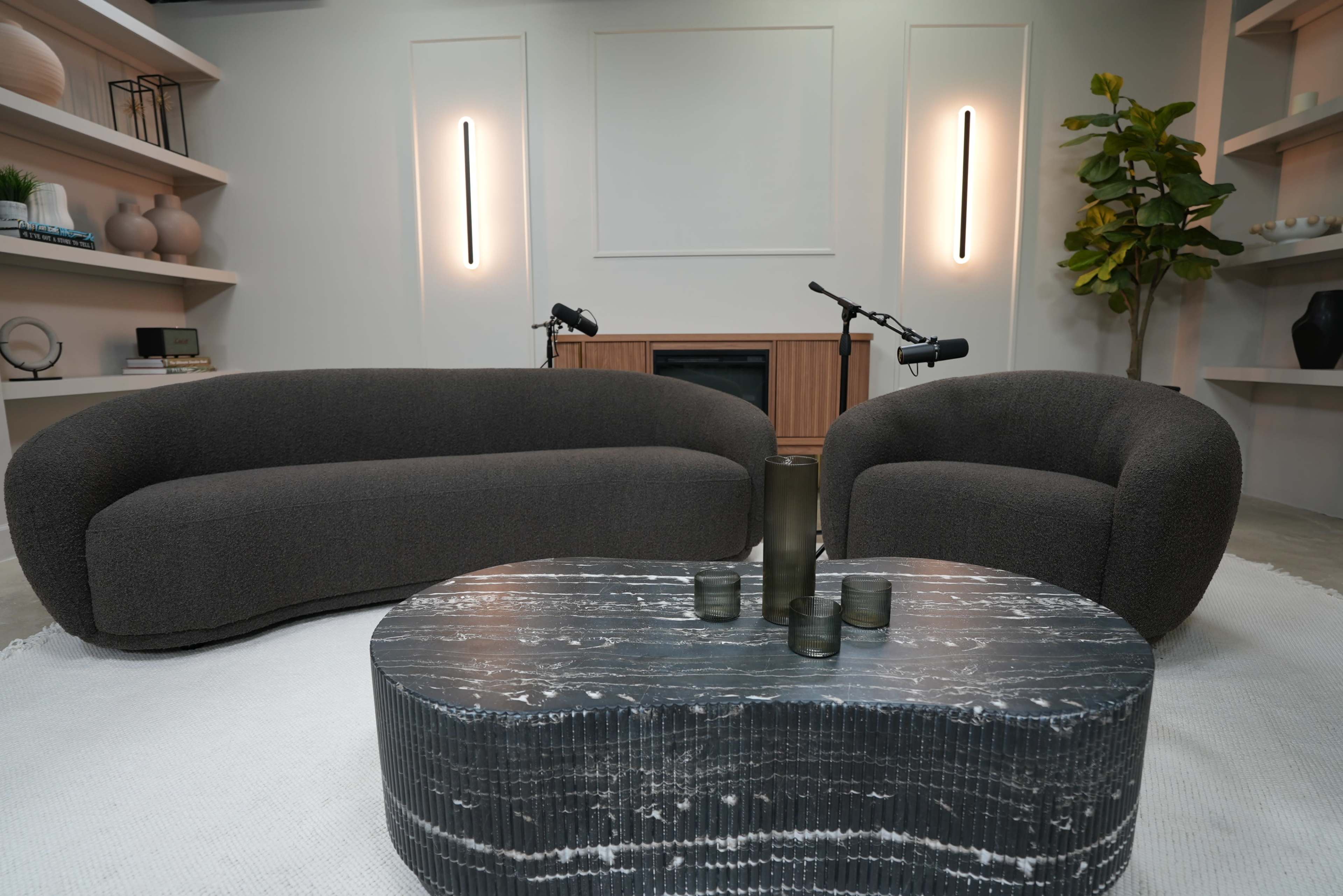 The image shows a modern living room setup featuring a large dark gray sofa and a matching round chair, positioned around a textured black coffee table, with soft lighting and decorative elements on the shelves.