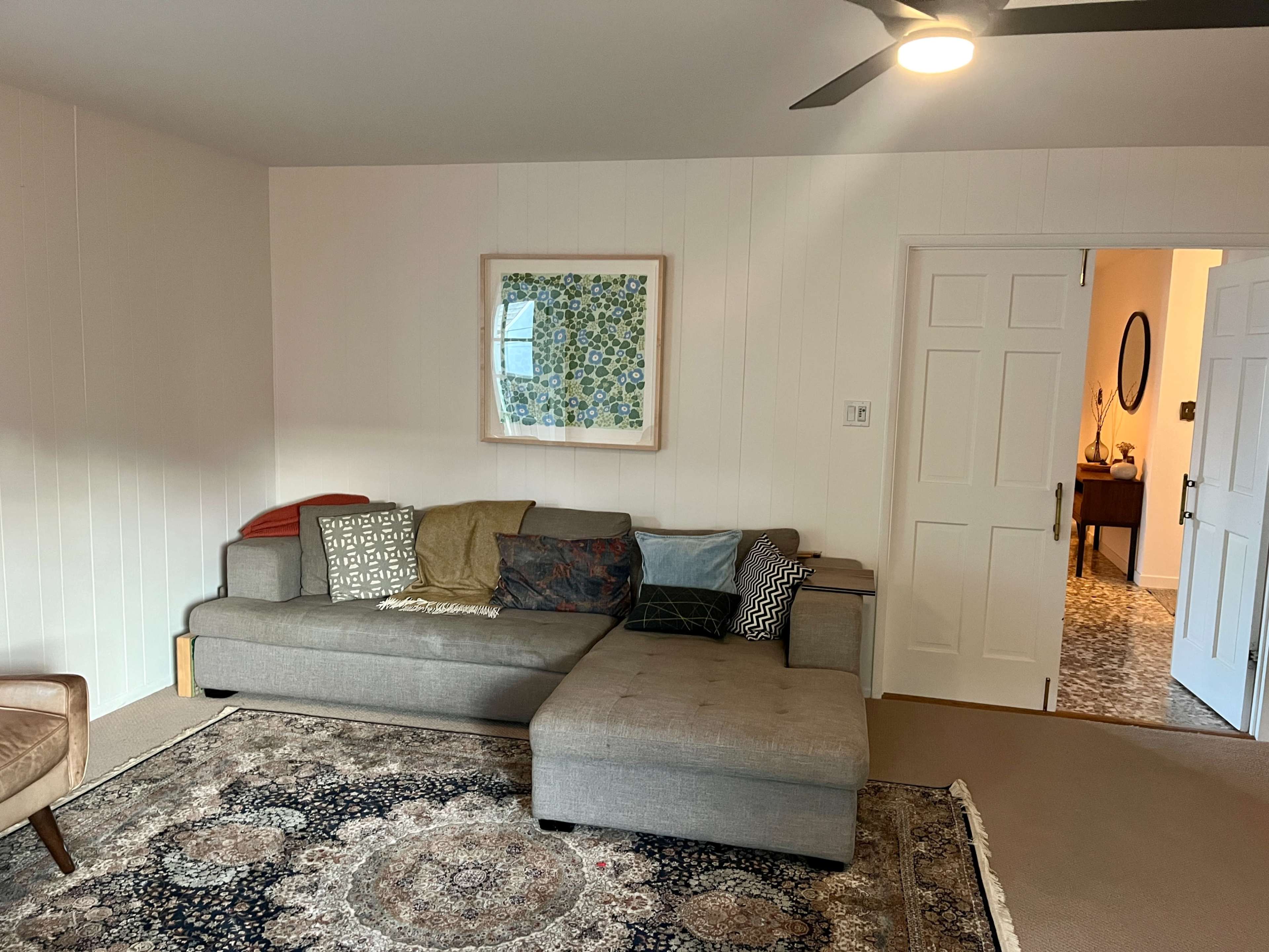 A living room with a sectional sofa, a patterned rug, and a framed piece of art on the wall, with a doorway leading to another room in the background.