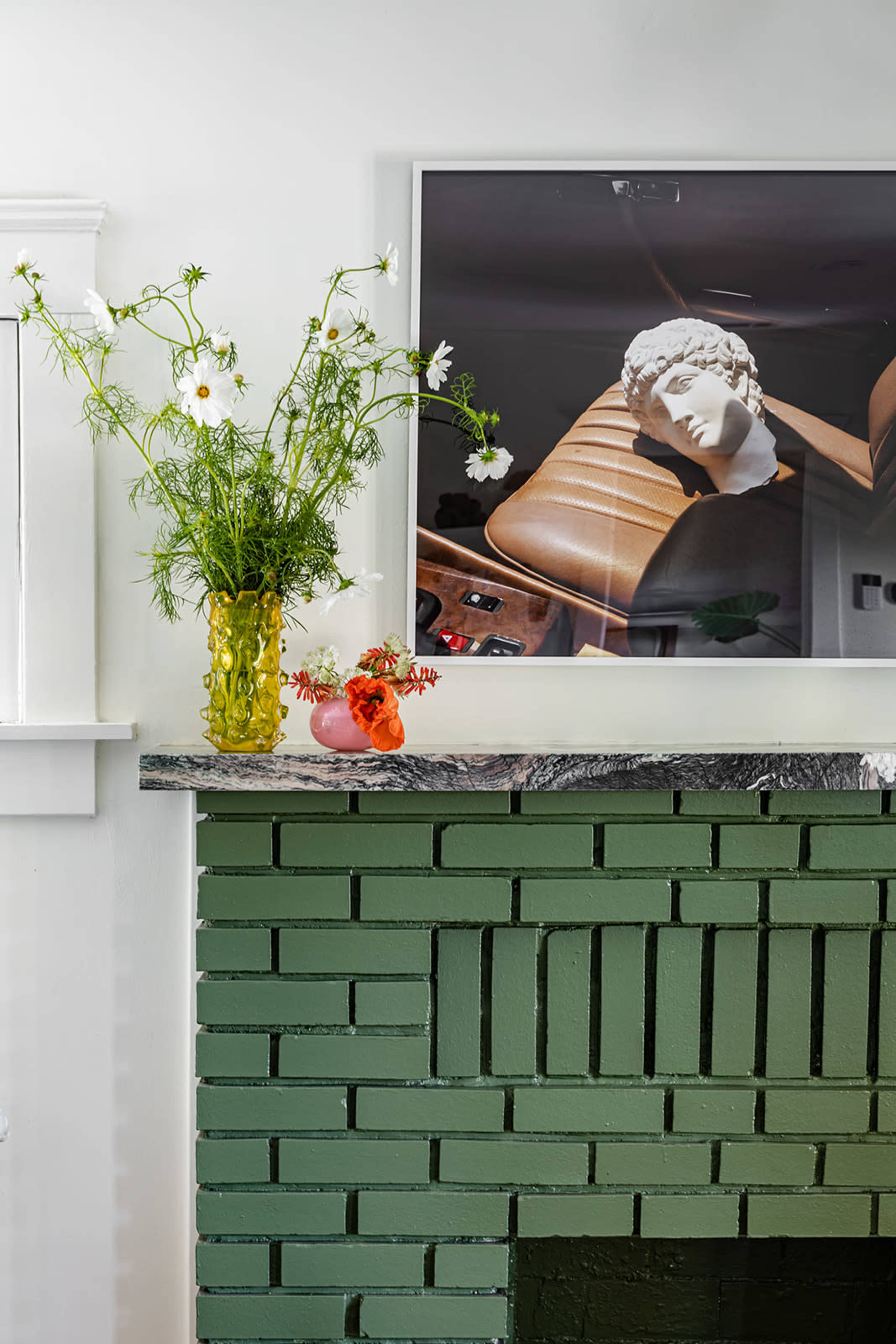 A green brick fireplace features a marble mantle adorned with a vase of flowers and a photograph of a classical bust on the wall above.