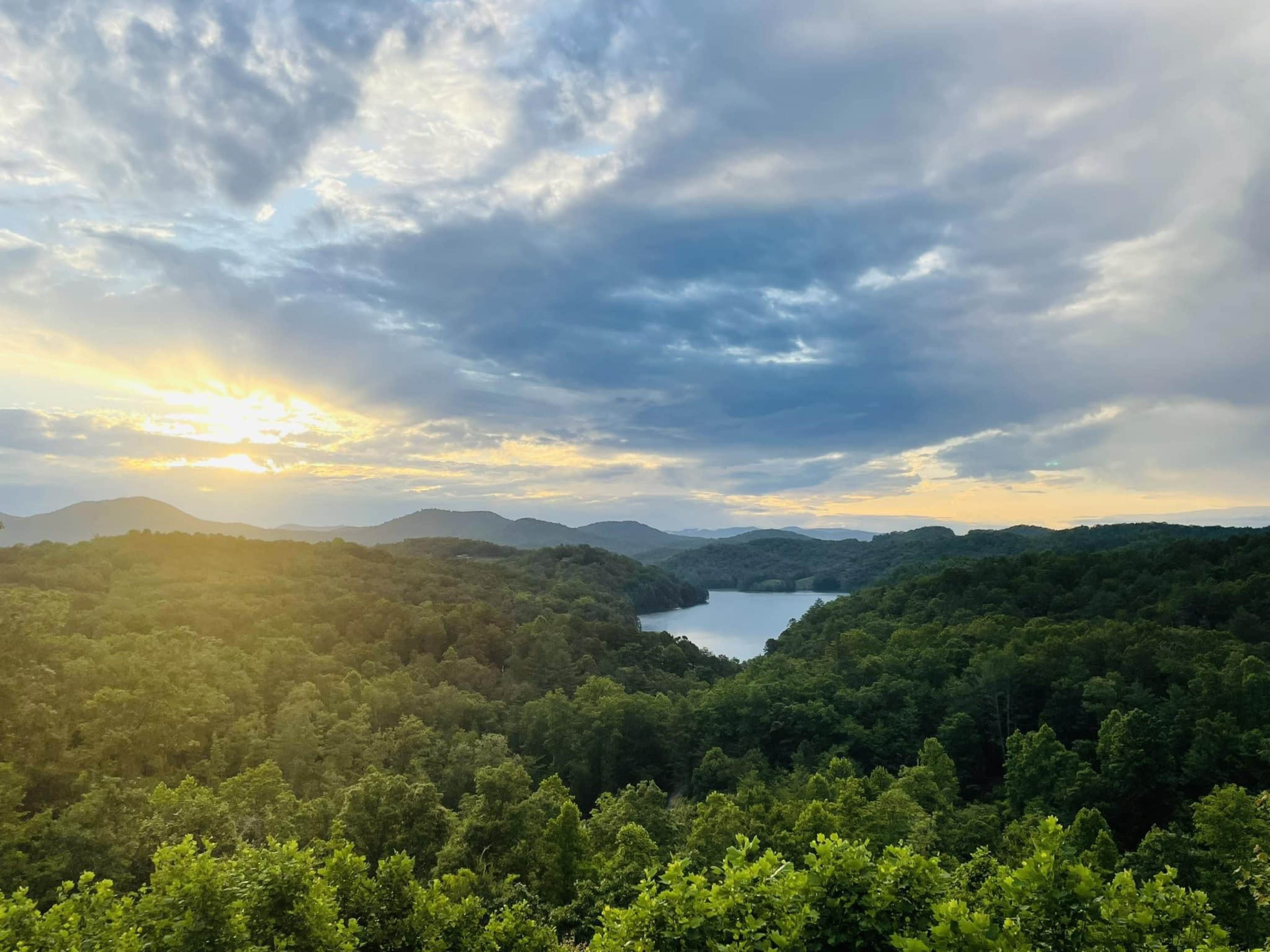 A serene landscape features a lake surrounded by hills, with the sun setting behind the mountains and clouds.