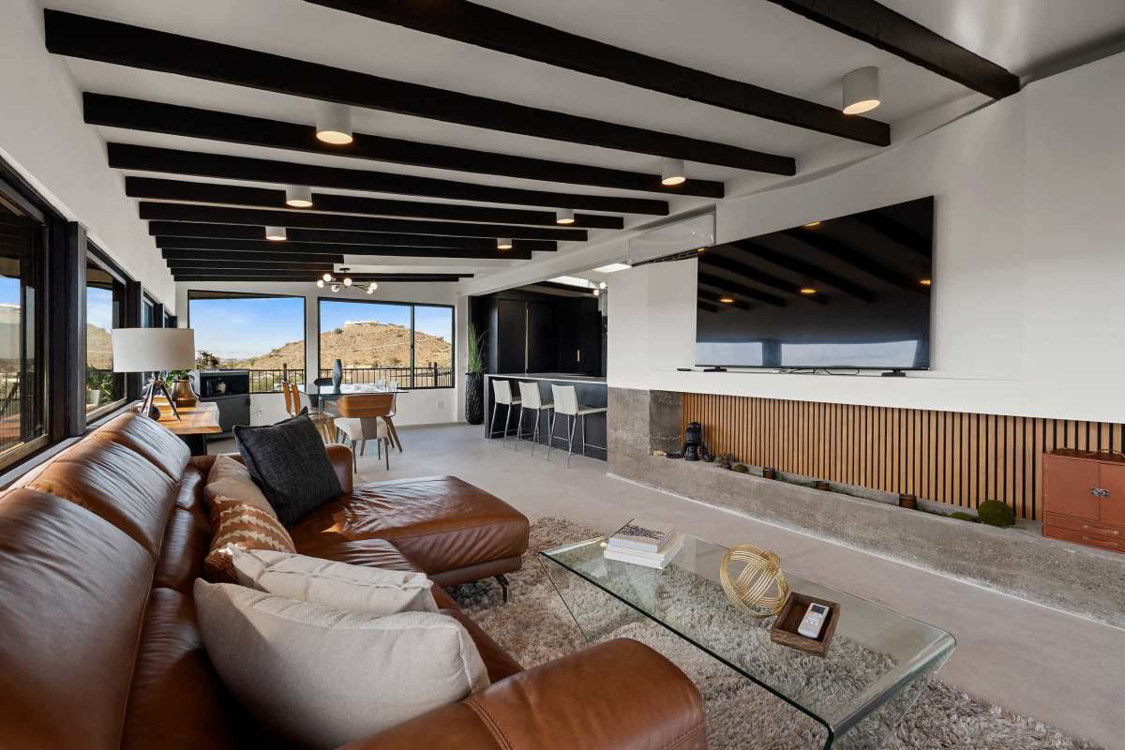 The image shows a modern living room featuring a brown leather sofa, a glass coffee table, and large windows that provide a view of a mountainous landscape.