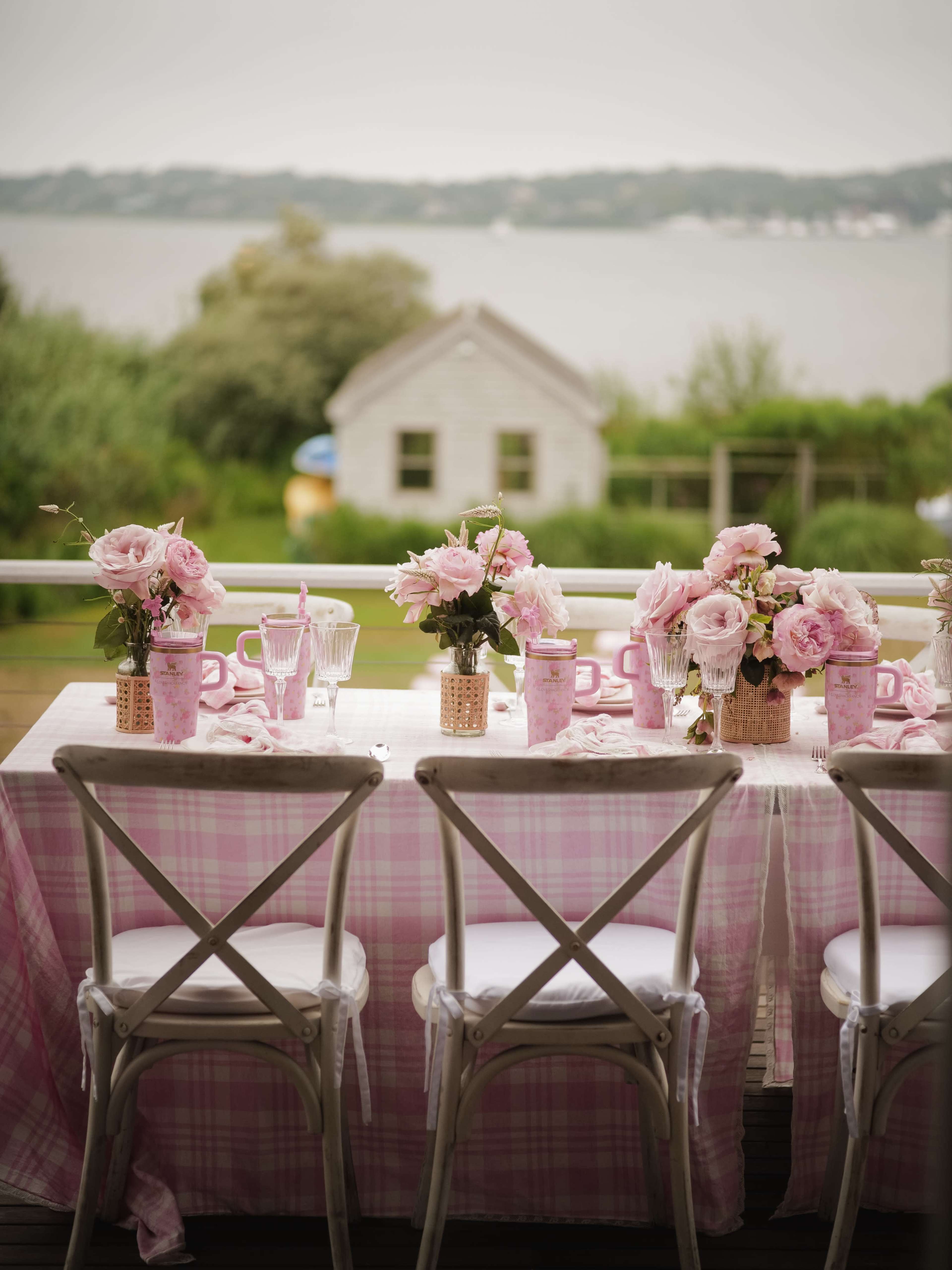 A long table is set with pink checkered tablecloths and floral centerpieces overlooking a body of water and a small shed in the background.