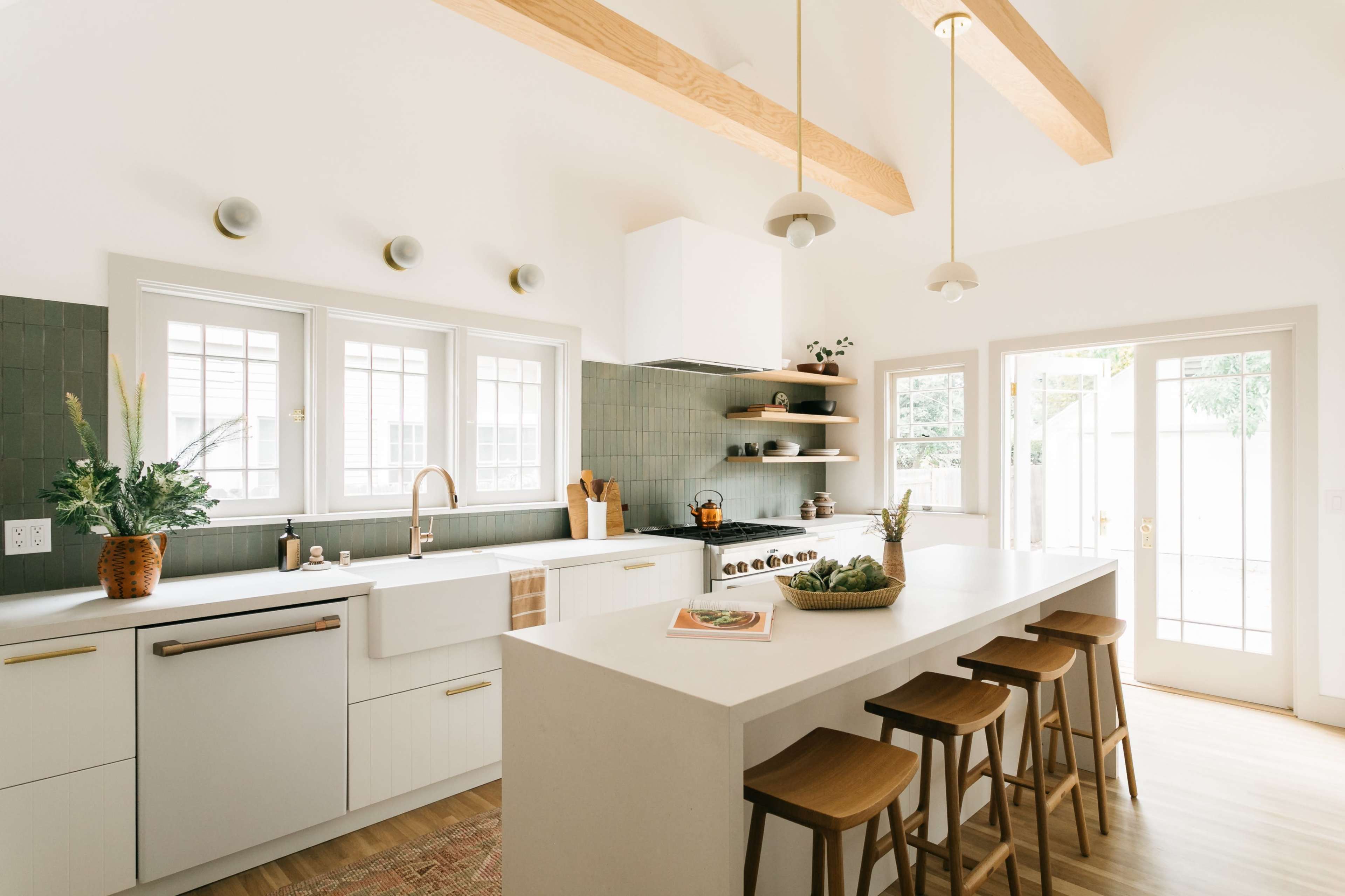 A modern kitchen features a large island with seating, light wood beams on the ceiling, and green tile backsplash behind a white sink and stove.