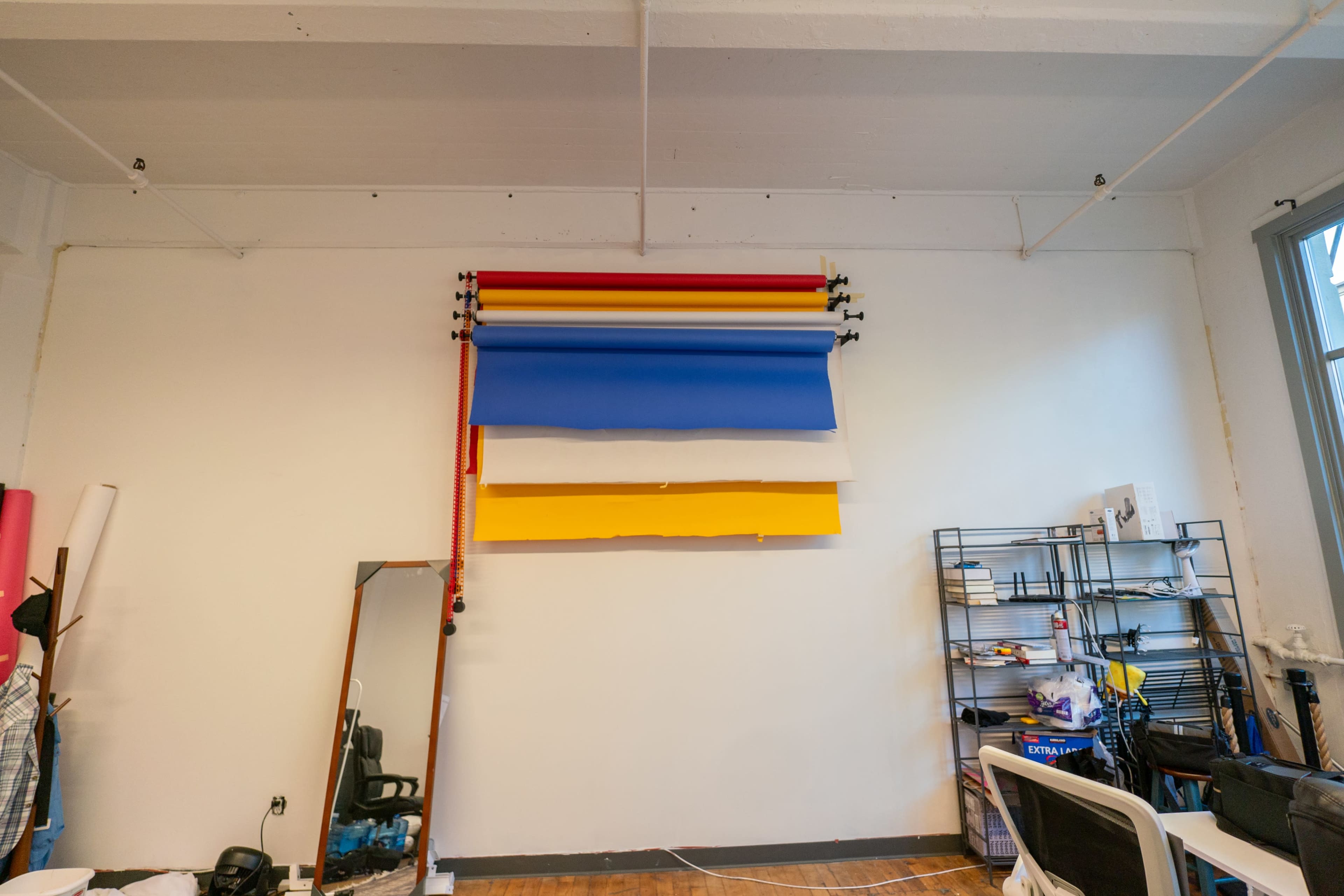 A wall with several rolls of colored backdrop paper hanging from hooks, alongside a mirror and shelves with various items.
