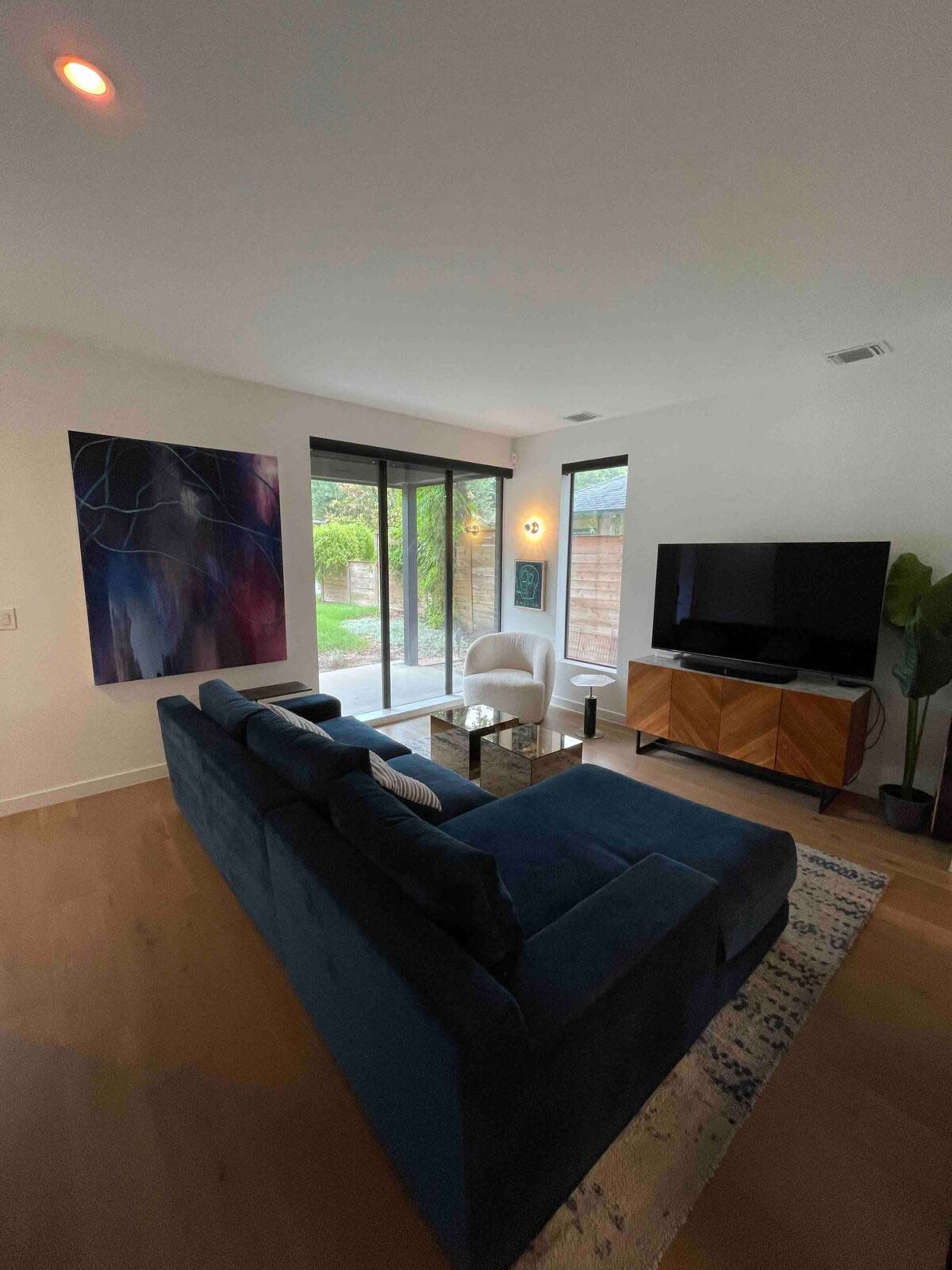The image shows a modern living room with a blue sofa, a white chair, a television, and large windows allowing natural light to enter.