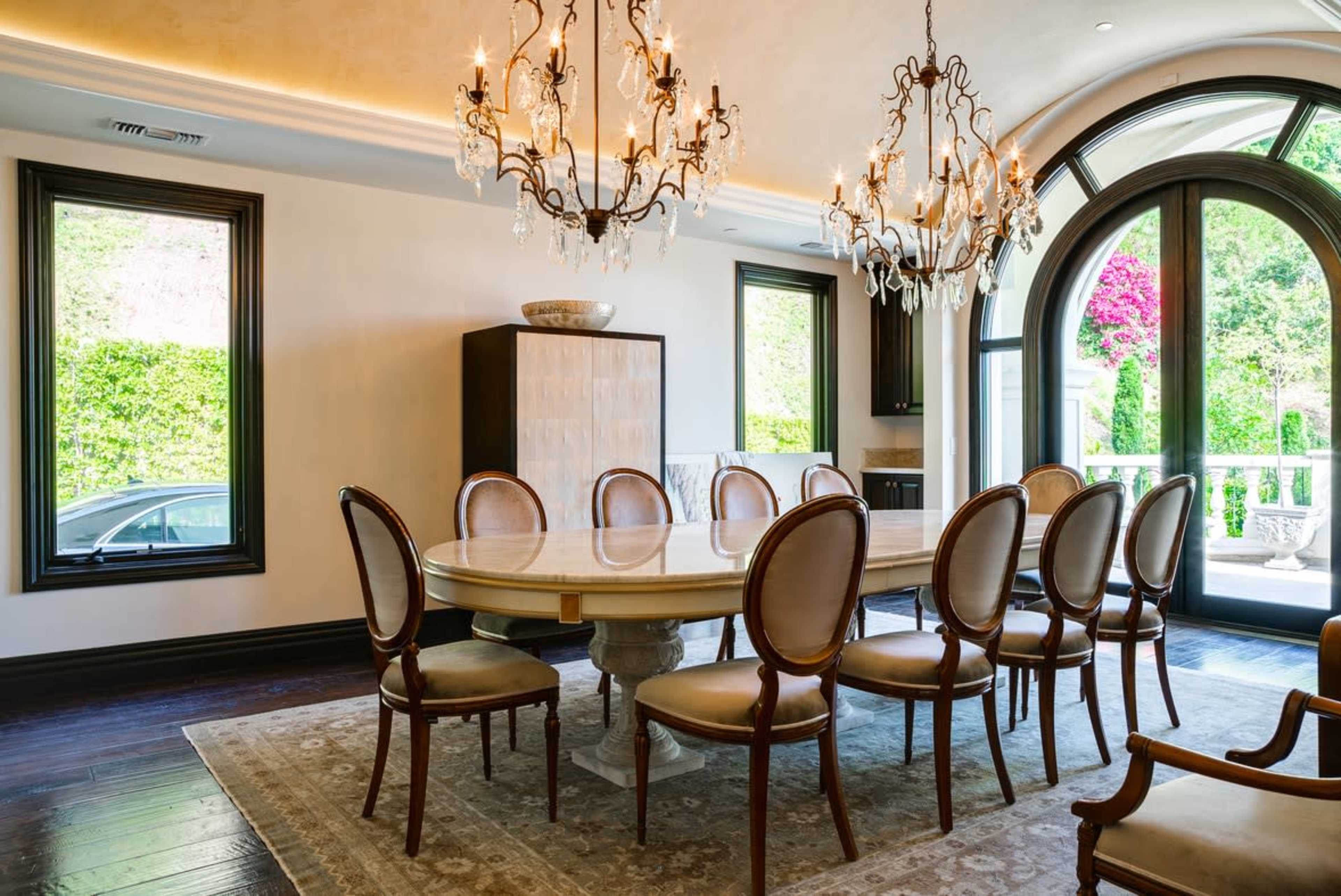 A spacious dining room features an oval table surrounded by chairs, decorative chandeliers overhead, and large arched windows with greenery visible outside.