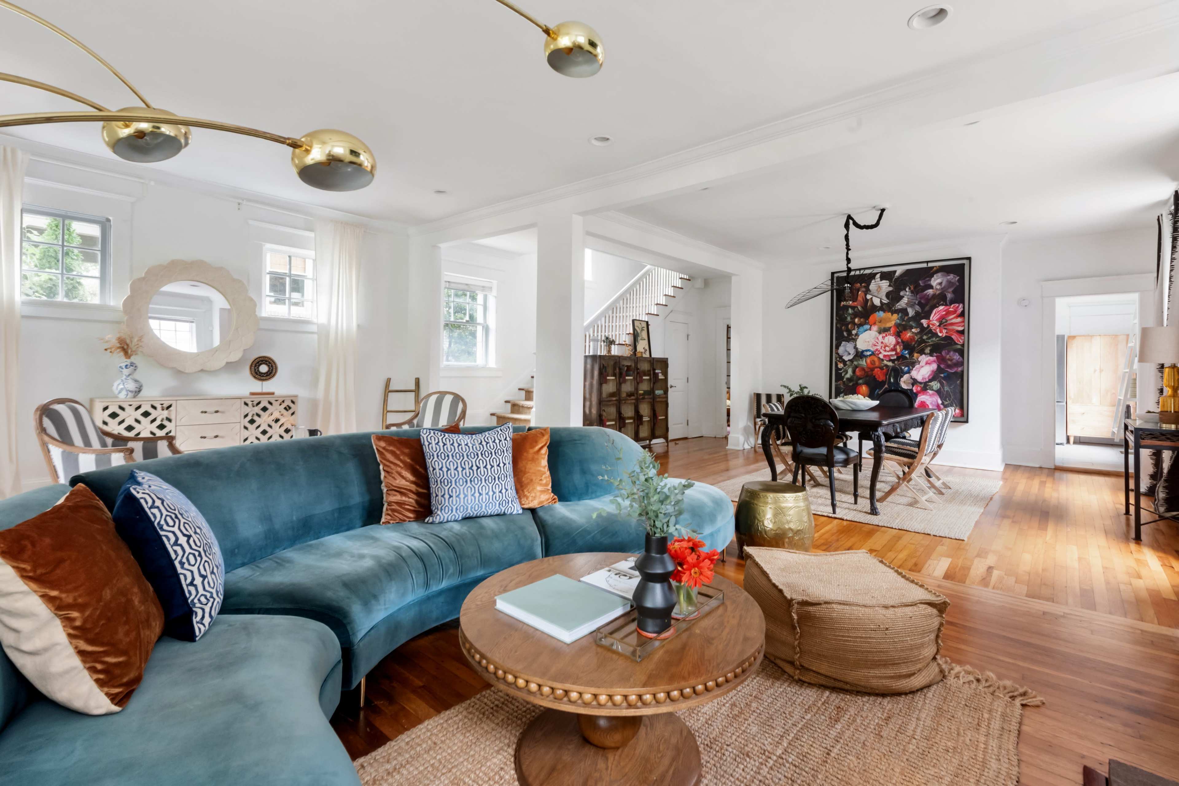 The living room features a modern blue sectional sofa, a round coffee table with decorative accents, and a dining area with a large floral artwork on the wall.