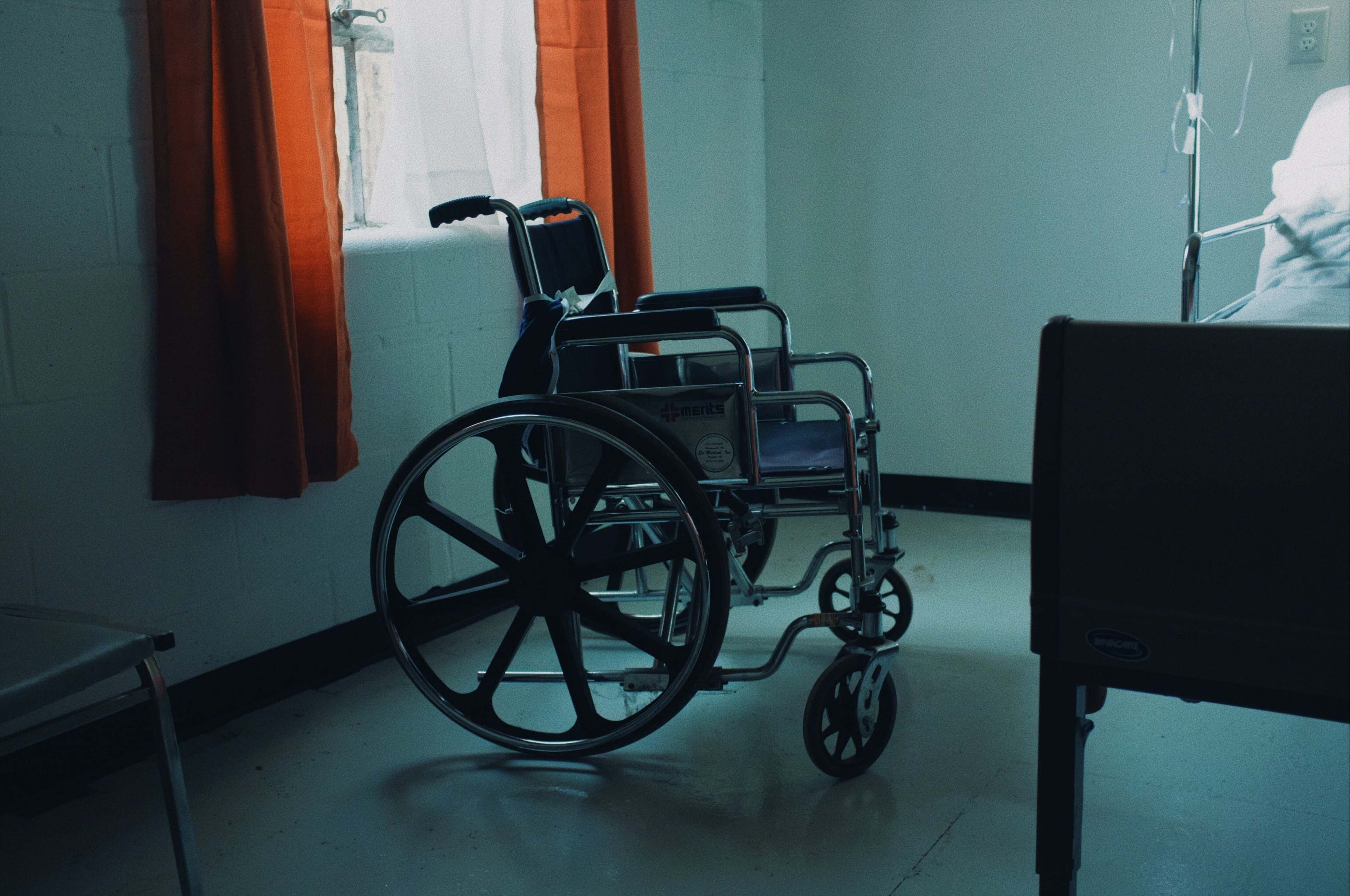 A wheelchair is positioned near a hospital bed in a dimly lit room with orange curtains.
