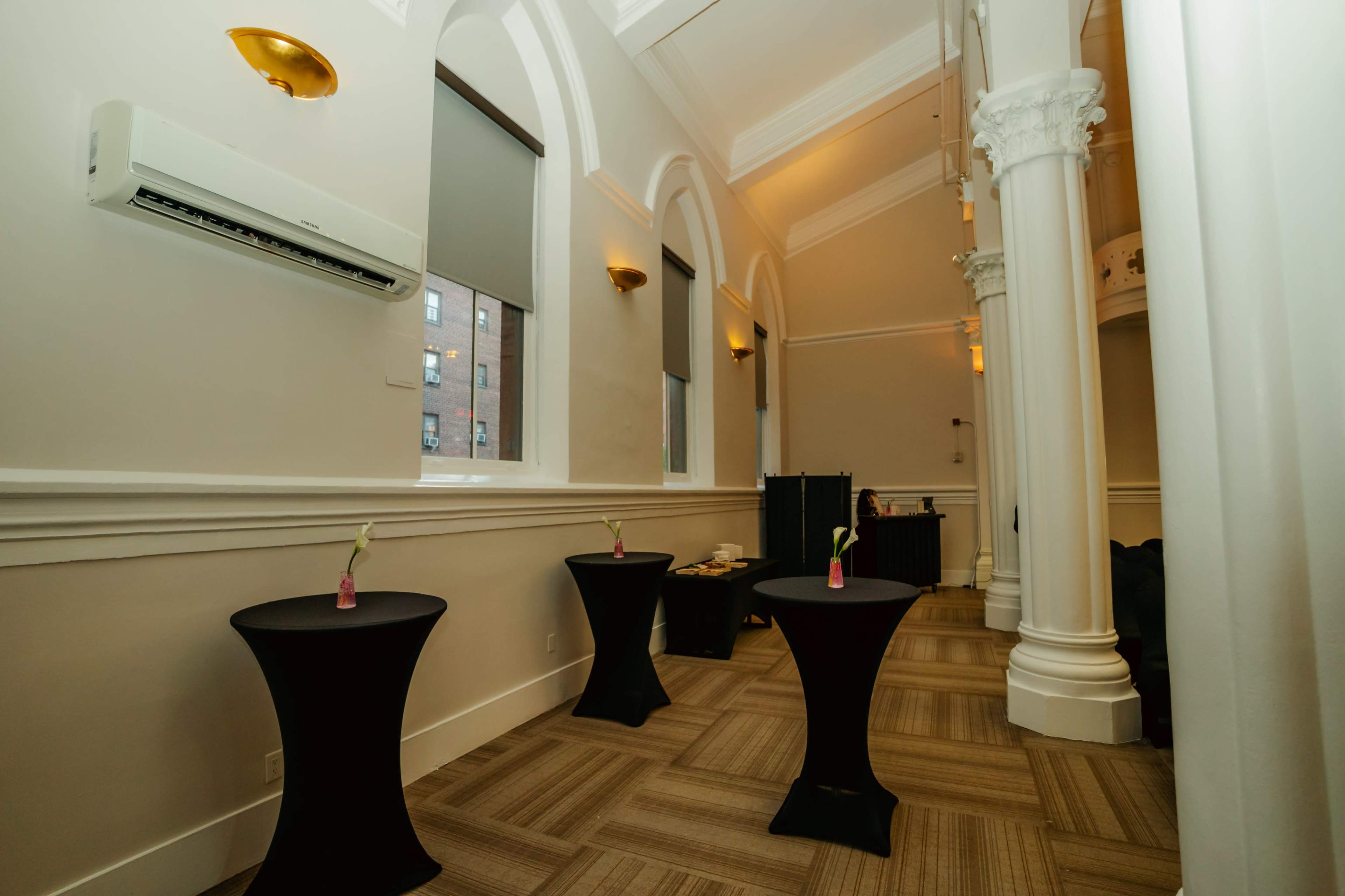 The image shows a spacious room with tall ceilings, featuring three black cocktail tables with small floral arrangements on them and wall-mounted lighting.
