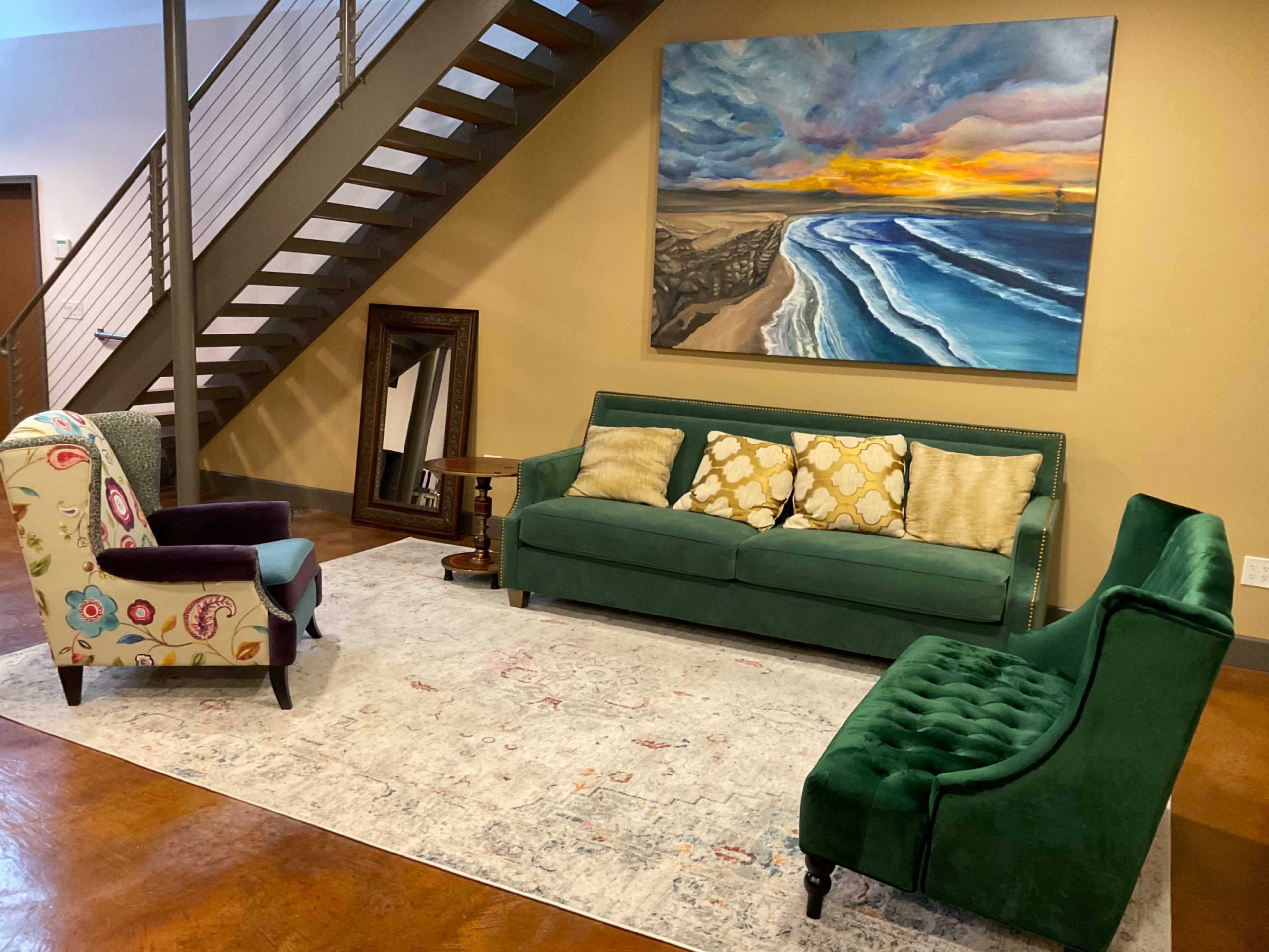 A living room with a green sofa, two accent chairs, a round table, a large painting of a seascape, and a staircase in the background.